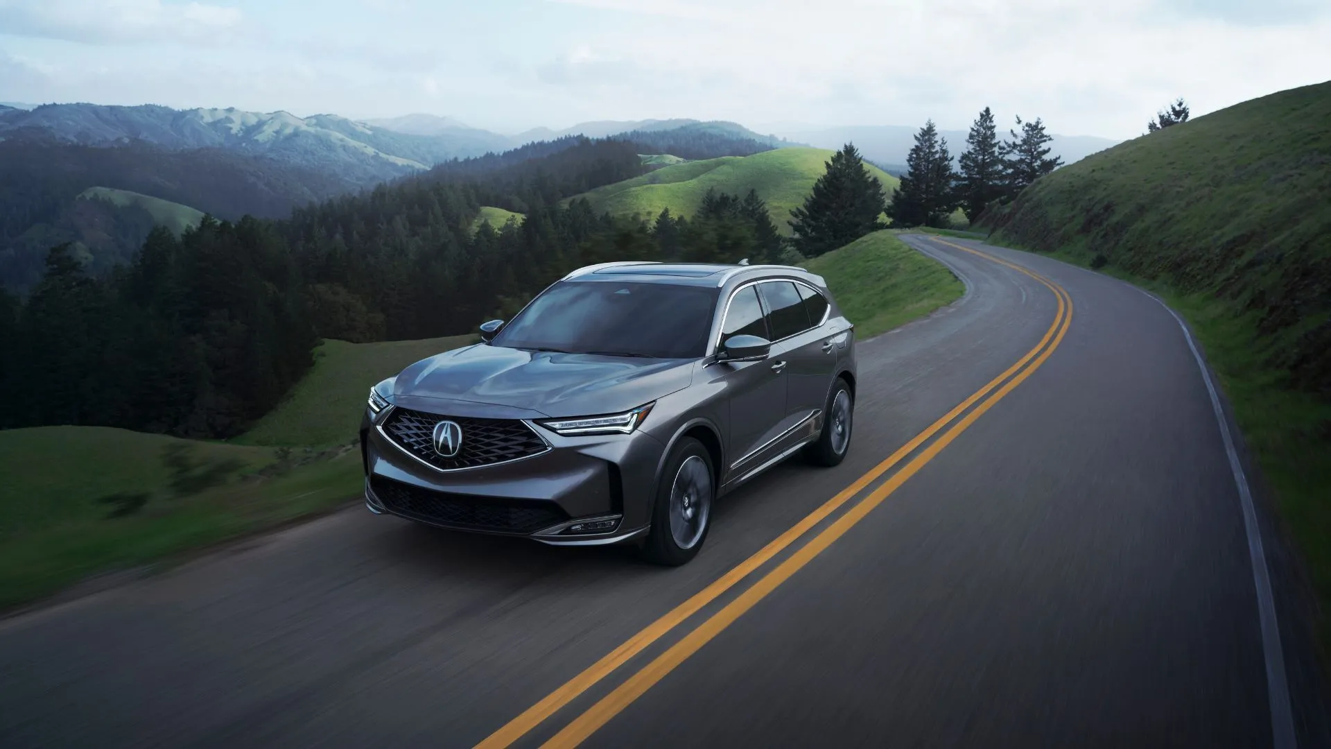Acura MDX 2025 màu xanh đậm đang chạy trên con đường nông thôn với núi non làm nền, thể hiện phong cách SUV hạng sang
