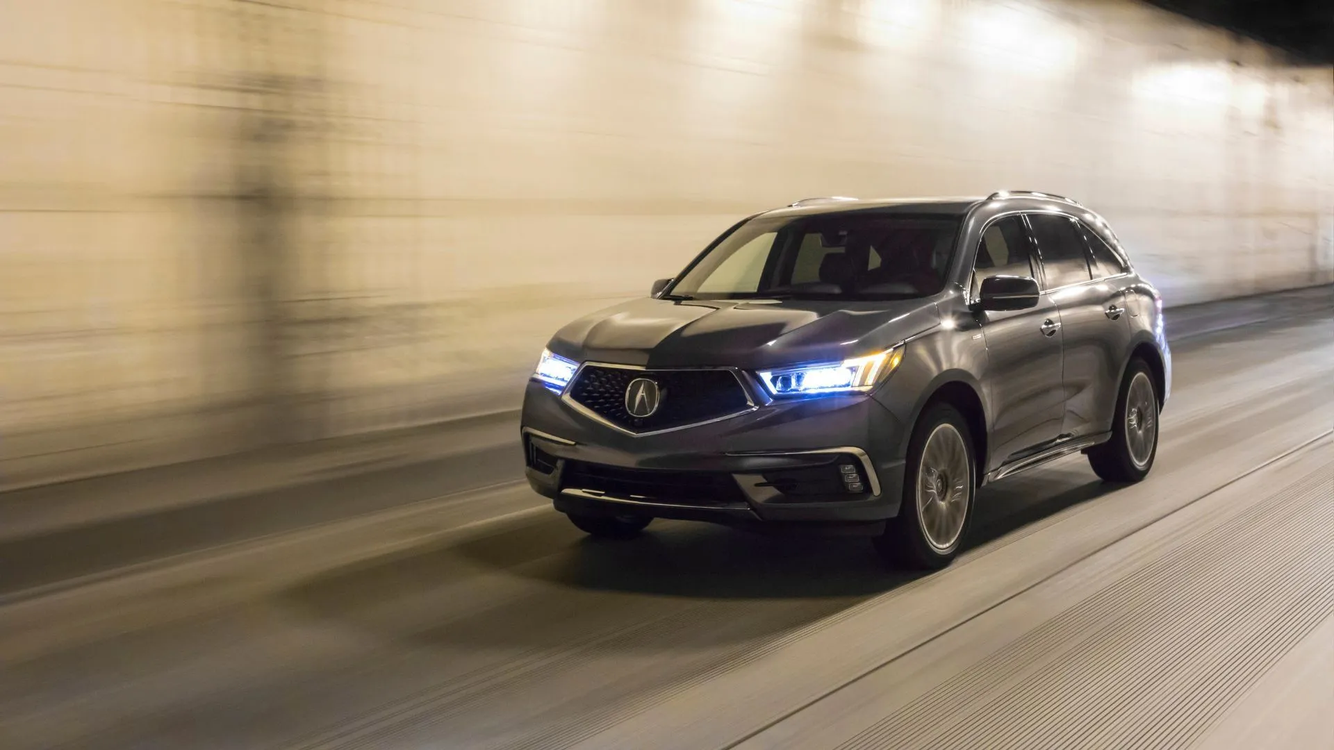 Acura MDX Sport Hybrid 2020 màu xám đang di chuyển qua hầm với đèn pha sáng, nhấn mạnh tầm quan trọng của hệ thống đèn hoạt động tốt.