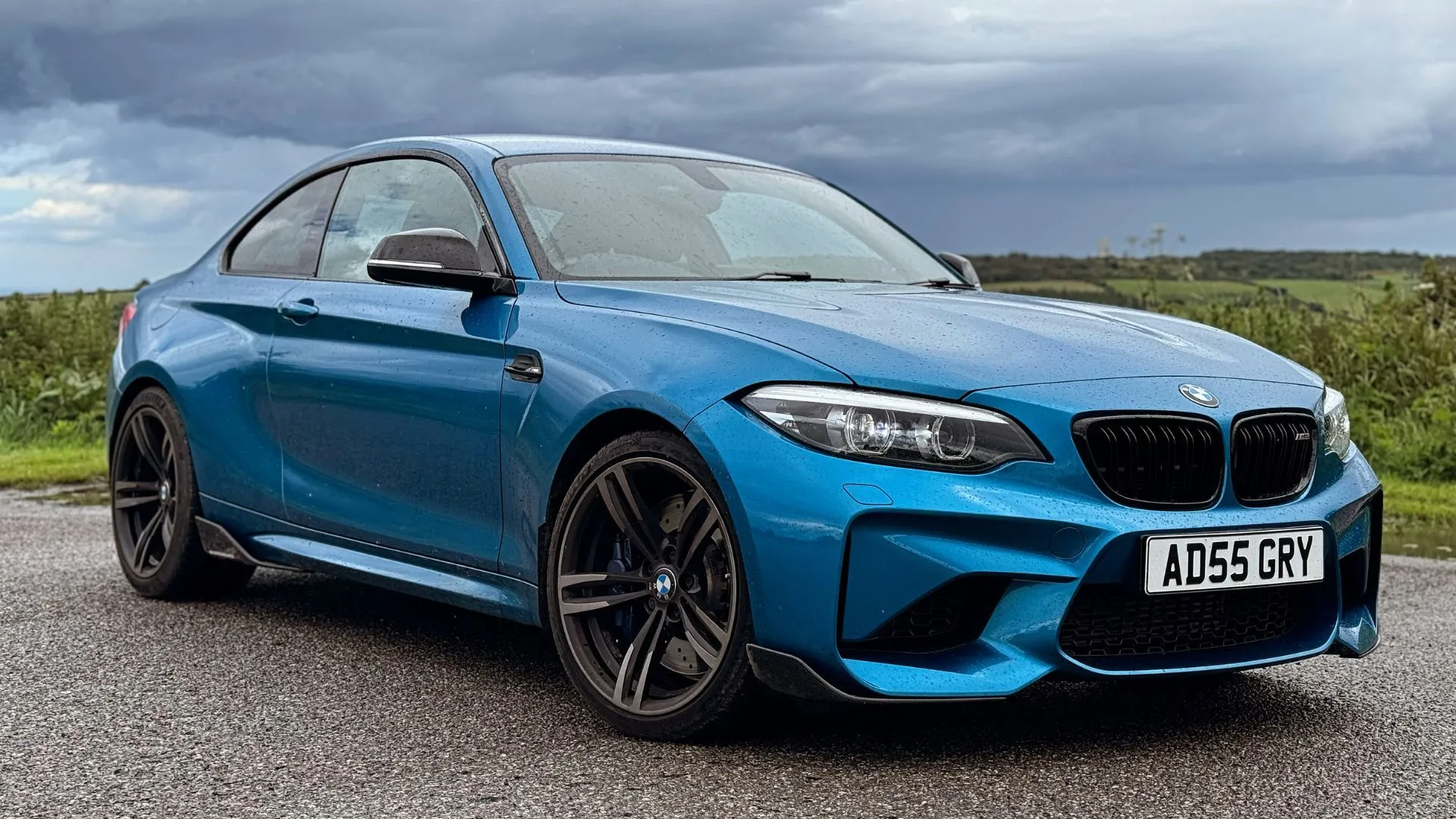 BMW M2 2018 màu xanh dương đậu tĩnh, nhìn từ góc 3/4 phía trước với cảnh đồng quê phía sau, thể hiện vẻ đẹp vượt thời gian.