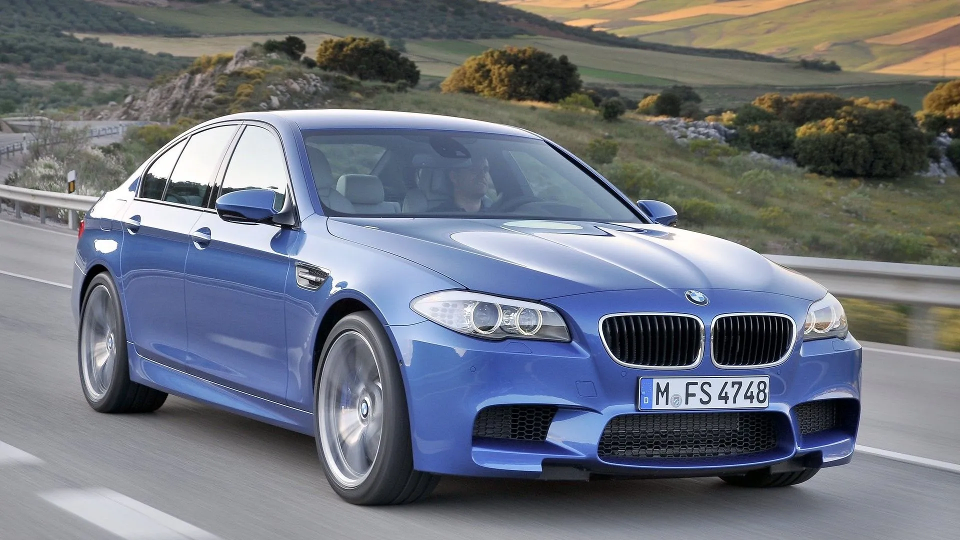 BMW M5 2013 màu xanh dương đang tăng tốc trên đường, góc nhìn 3/4 từ phía trước, thể hiện sự mạnh mẽ và tinh tế