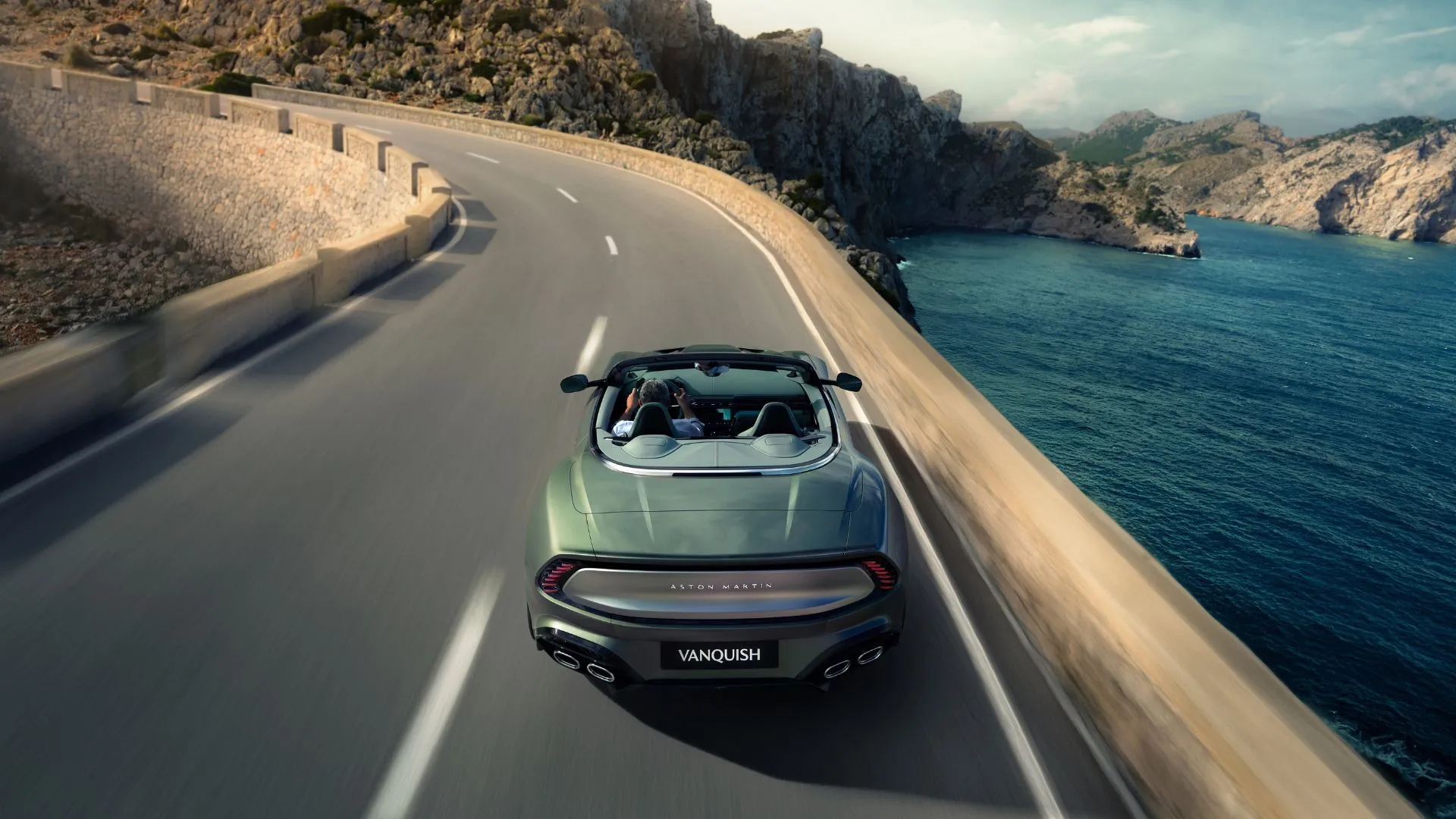 Cận cảnh phần đuôi xe Aston Martin Vanquish Volante màu bạc, mui trần đang mở, di chuyển trên con đường quanh co bên bờ hồ, nổi bật hệ thống đèn hậu và ống xả kép.