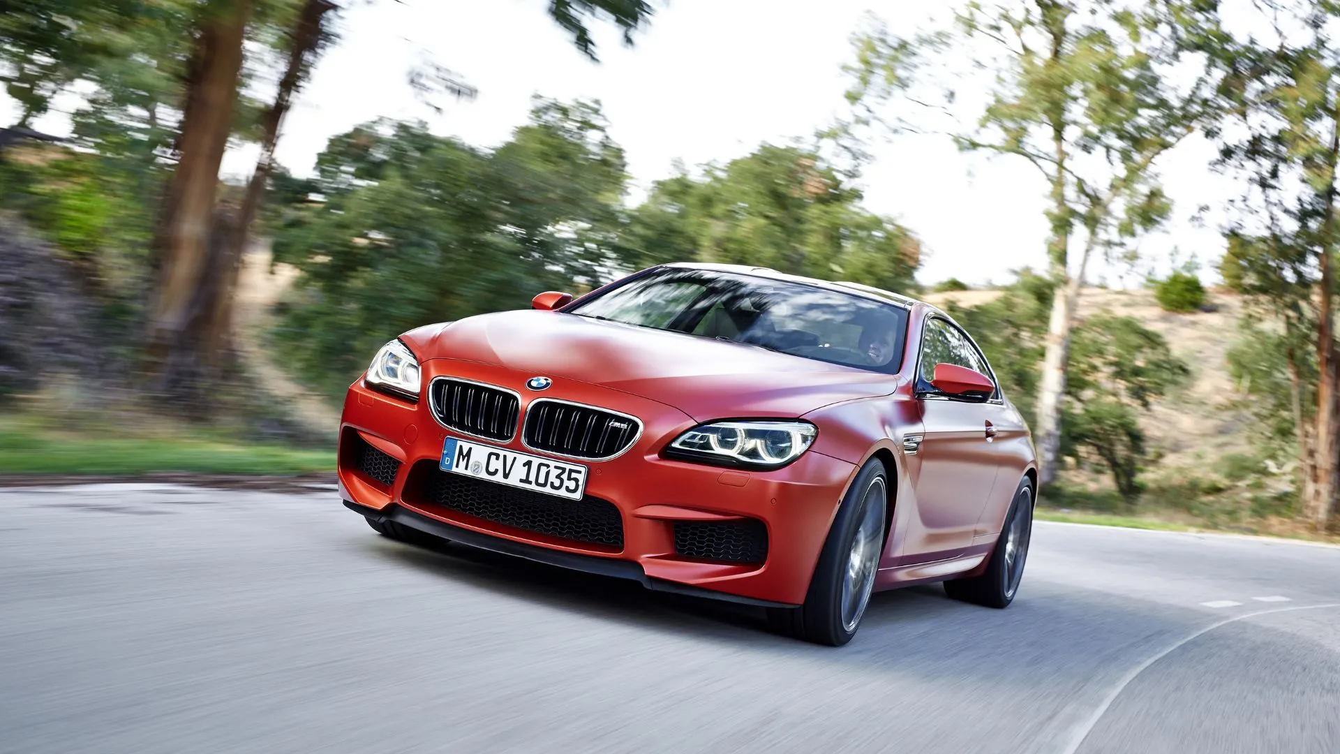 Chiếc BMW M6 Coupé màu cam đang chạy trên con đường quê quanh co, thể hiện sự kết hợp giữa hiệu suất và sang trọng.