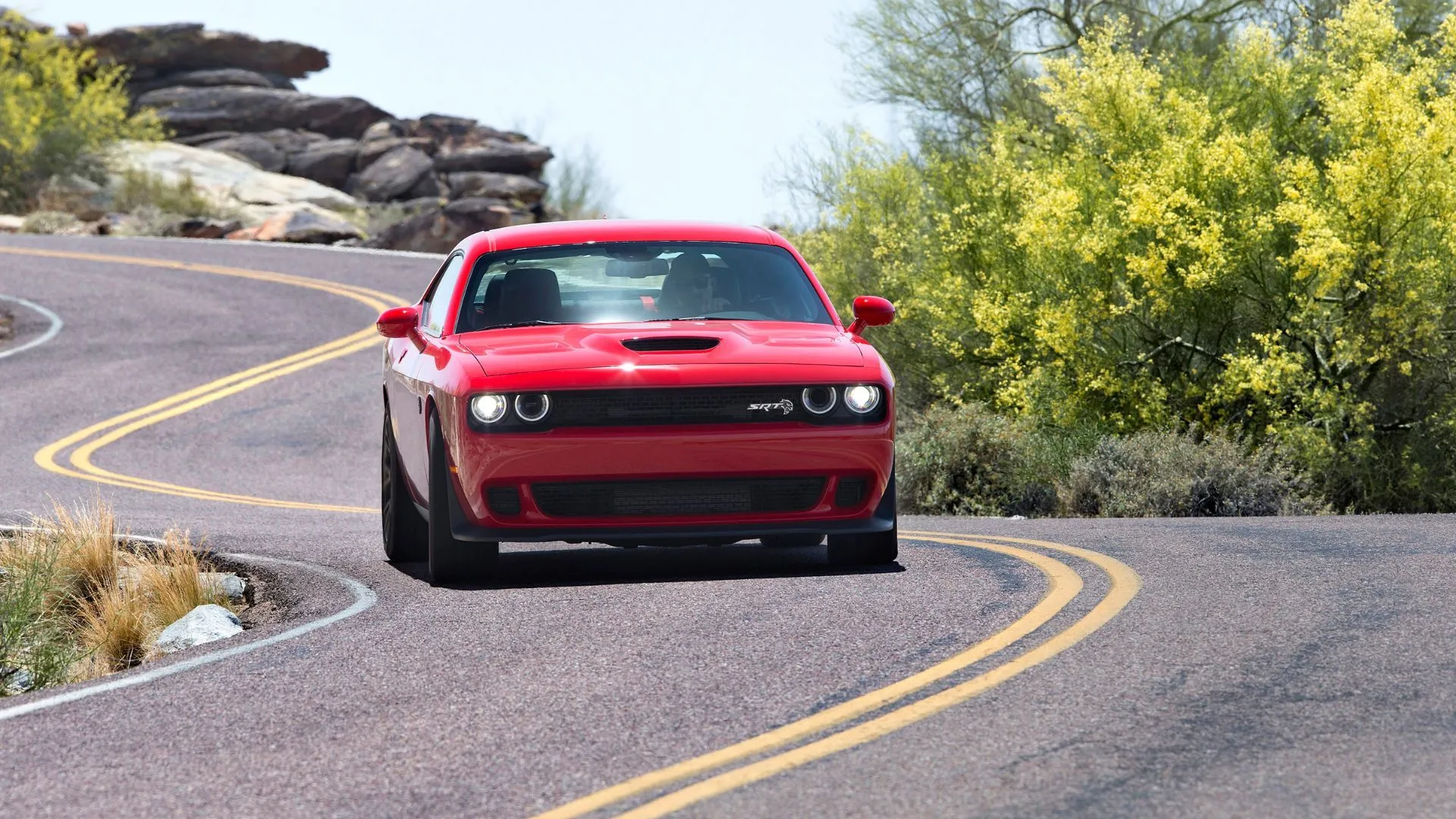 Chiếc Dodge Challenger SRT Hellcat màu đỏ đời 2018 đang chạy trên con đường quê quanh co, thể hiện sức mạnh cơ bắp và trải nghiệm lái đầy phấn khích.