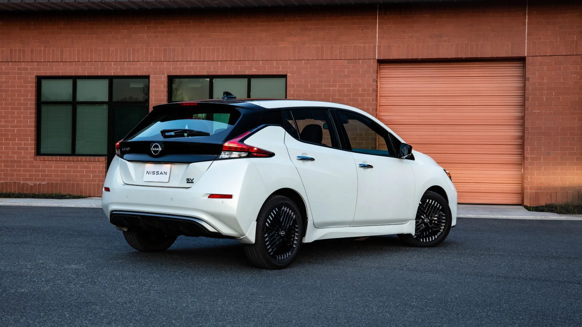 Chiếc Nissan Leaf 2025 màu trắng nhìn từ góc 3/4 phía sau đang đậu trên đường nhựa bên ngoài một tòa nhà gạch, cho thấy vẻ ngoài thực dụng và thân thiện môi trường.