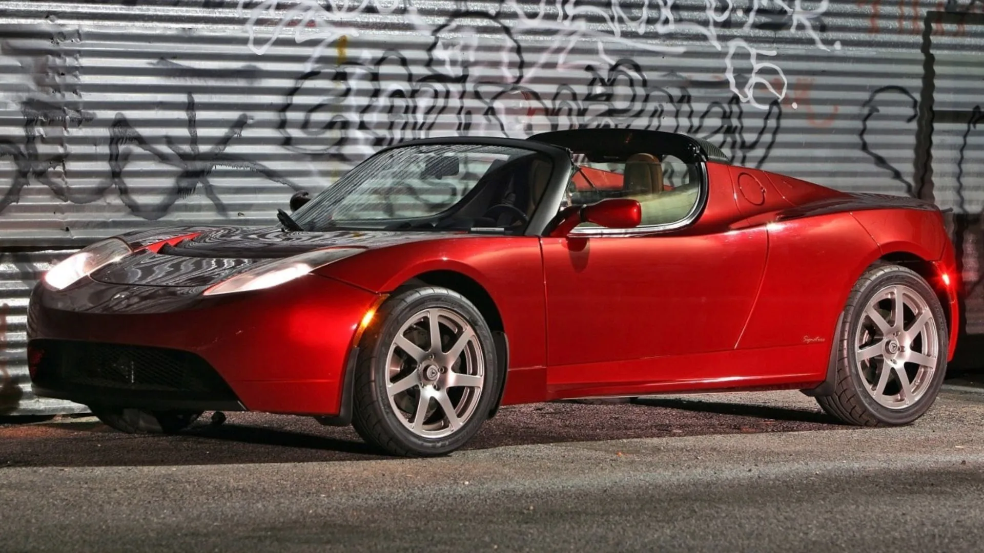 Chiếc Tesla Roadster thế hệ đầu tiên màu đỏ đậu trước cửa cuốn kim loại dán graffiti.