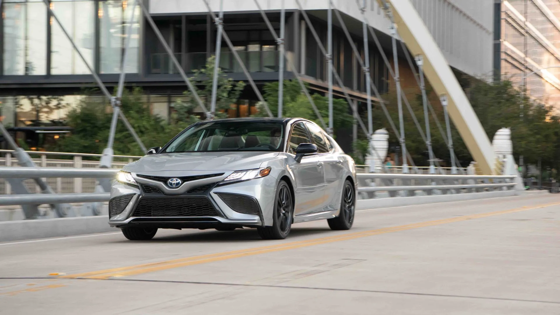 Chiếc Toyota Camry Hybrid 2022 màu bạc di chuyển trong thành phố, biểu tượng cho dòng xe hybrid được ưa chuộng.