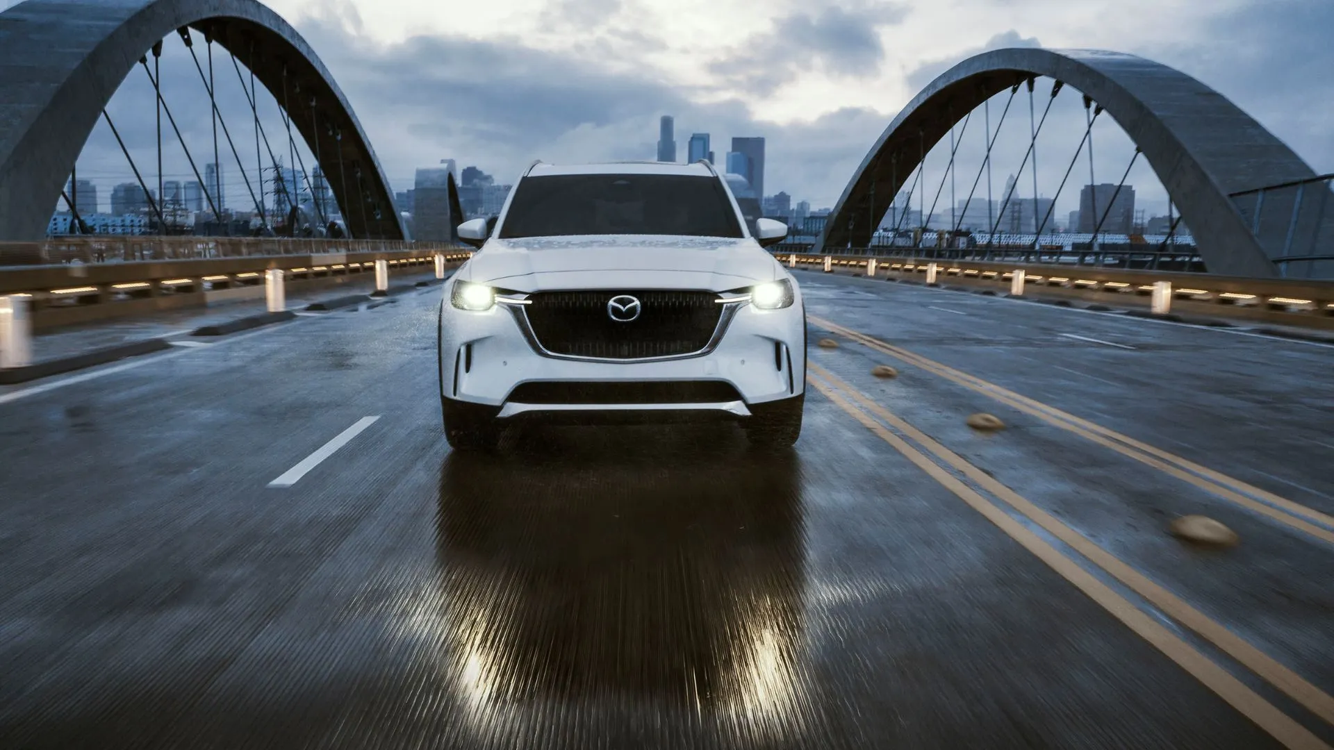 Đầu xe Mazda CX-90 2025 màu trắng đang lái qua cầu với khung cảnh thành phố phía sau.
