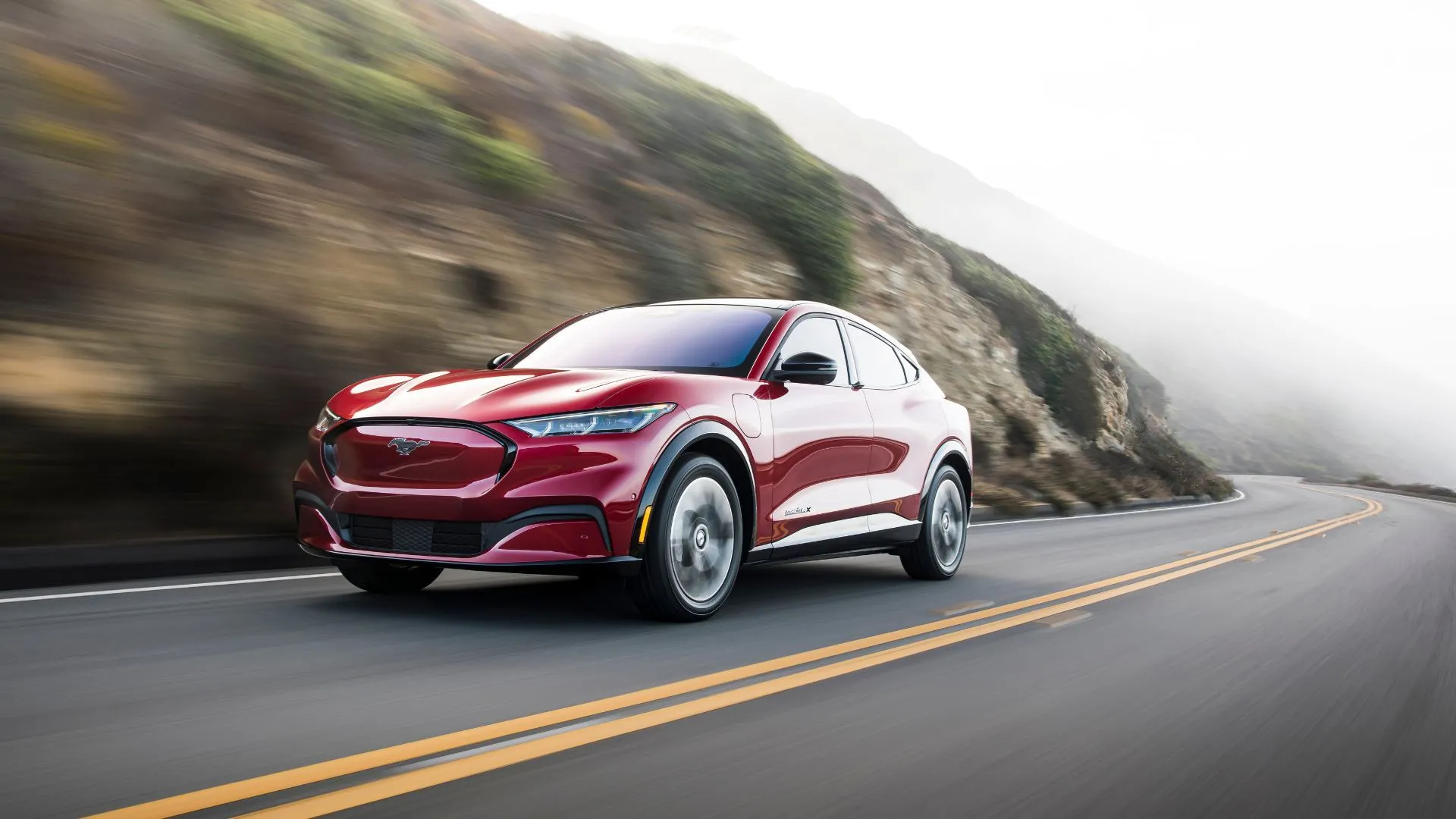 Ford Mustang Mach-E 2025 màu đỏ chạy trên đường quê uốn lượn, thể hiện khả năng vận hành linh hoạt và phạm vi hoạt động mở rộng.