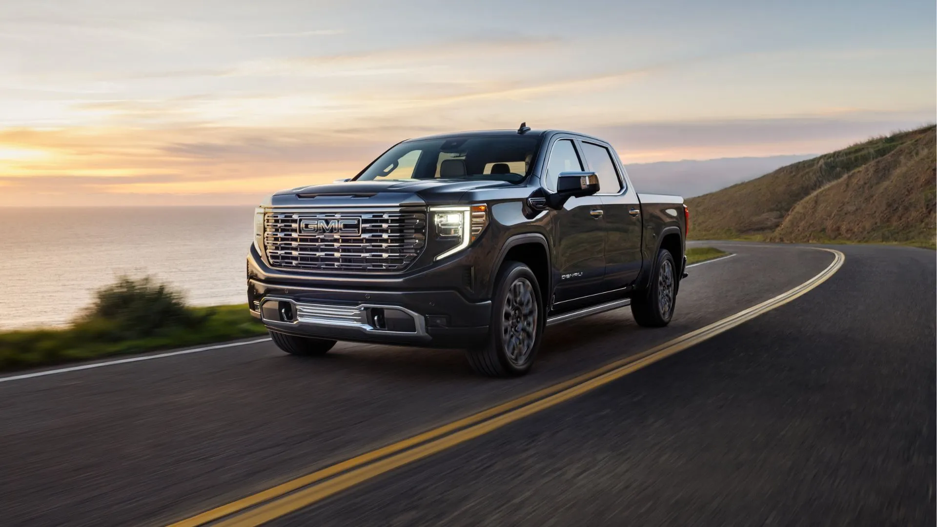 GMC Sierra Denali Ultimate 2025 màu xám bạc đang di chuyển trên đường cạnh bờ biển, minh họa cho dòng xe sử dụng động cơ V8 của GM