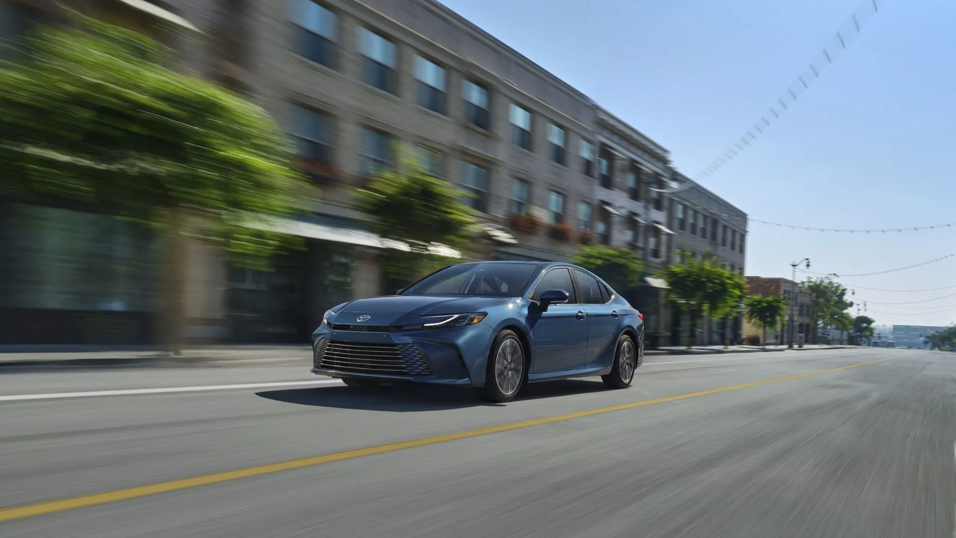Góc 3/4 phía trước của xe sedan Toyota Camry Hybrid LE 2025 màu xanh dương đang lái qua thành phố.