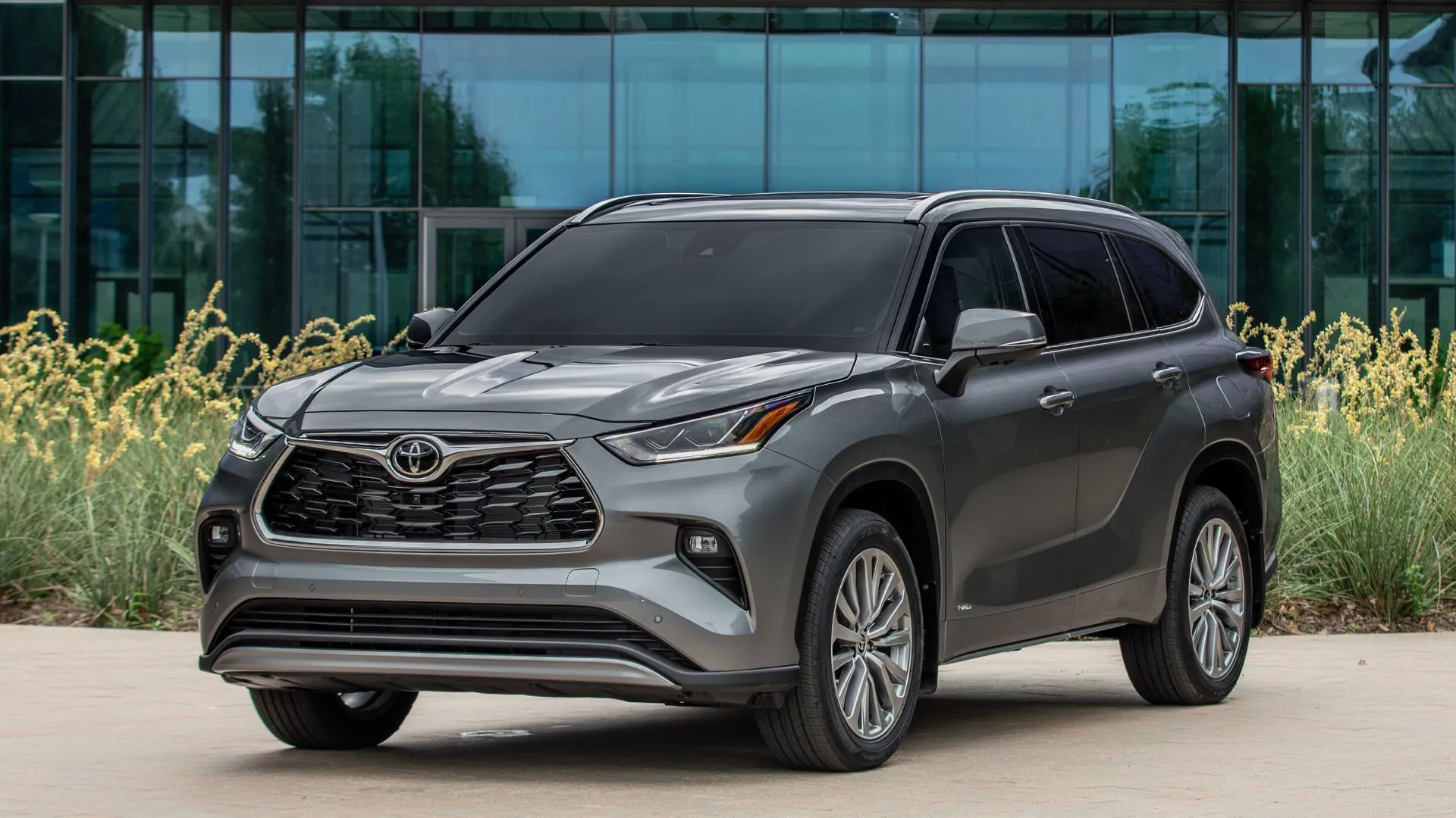 Góc 3/4 phía trước xe Toyota Highlander Hybrid 2025 màu xám đậu trước tòa nhà kính