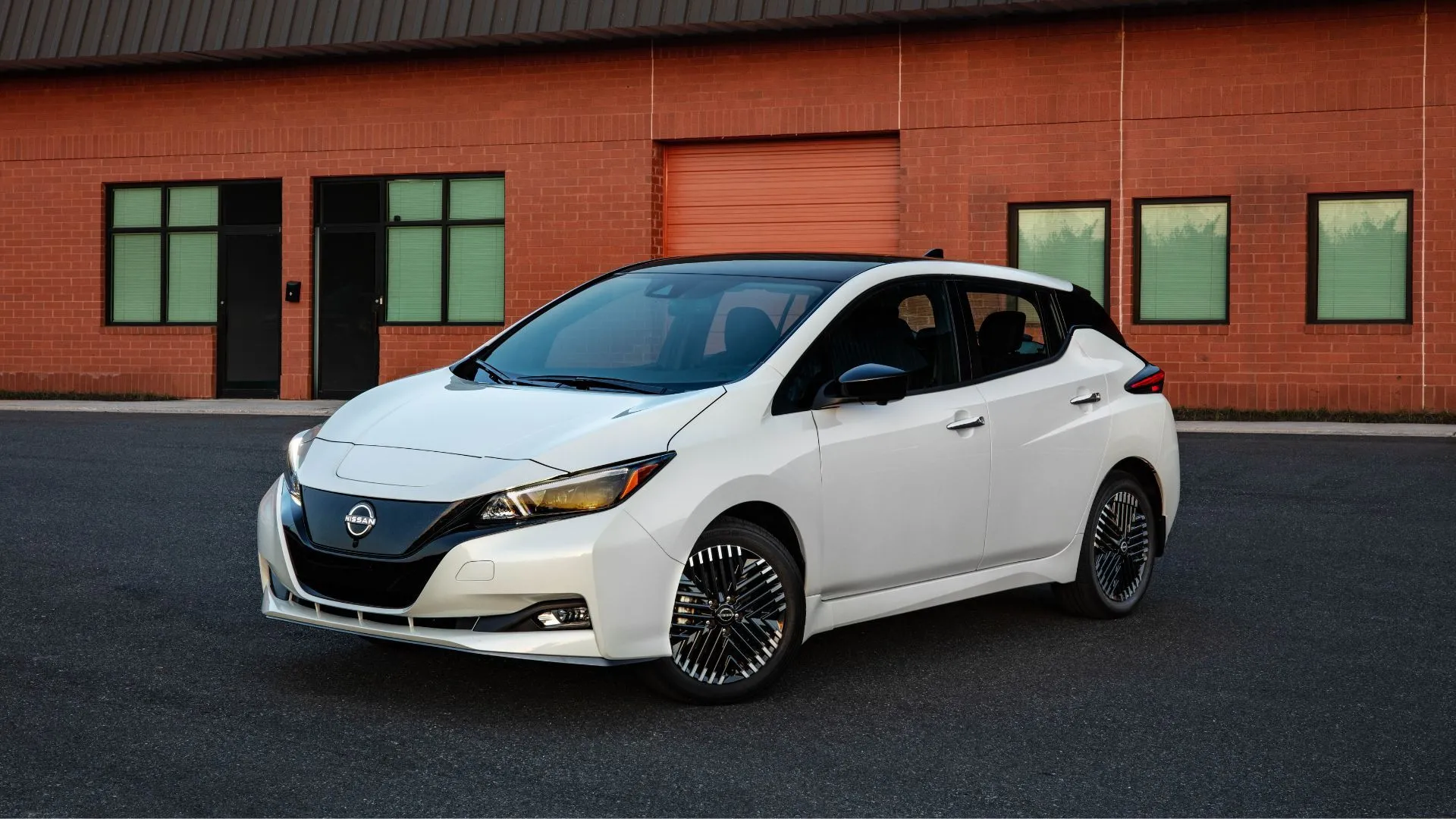 Góc nhìn ba phần tư phía trước của một chiếc Nissan Leaf 2025 màu trắng đỗ trên mặt đường bên ngoài một tòa nhà gạch.