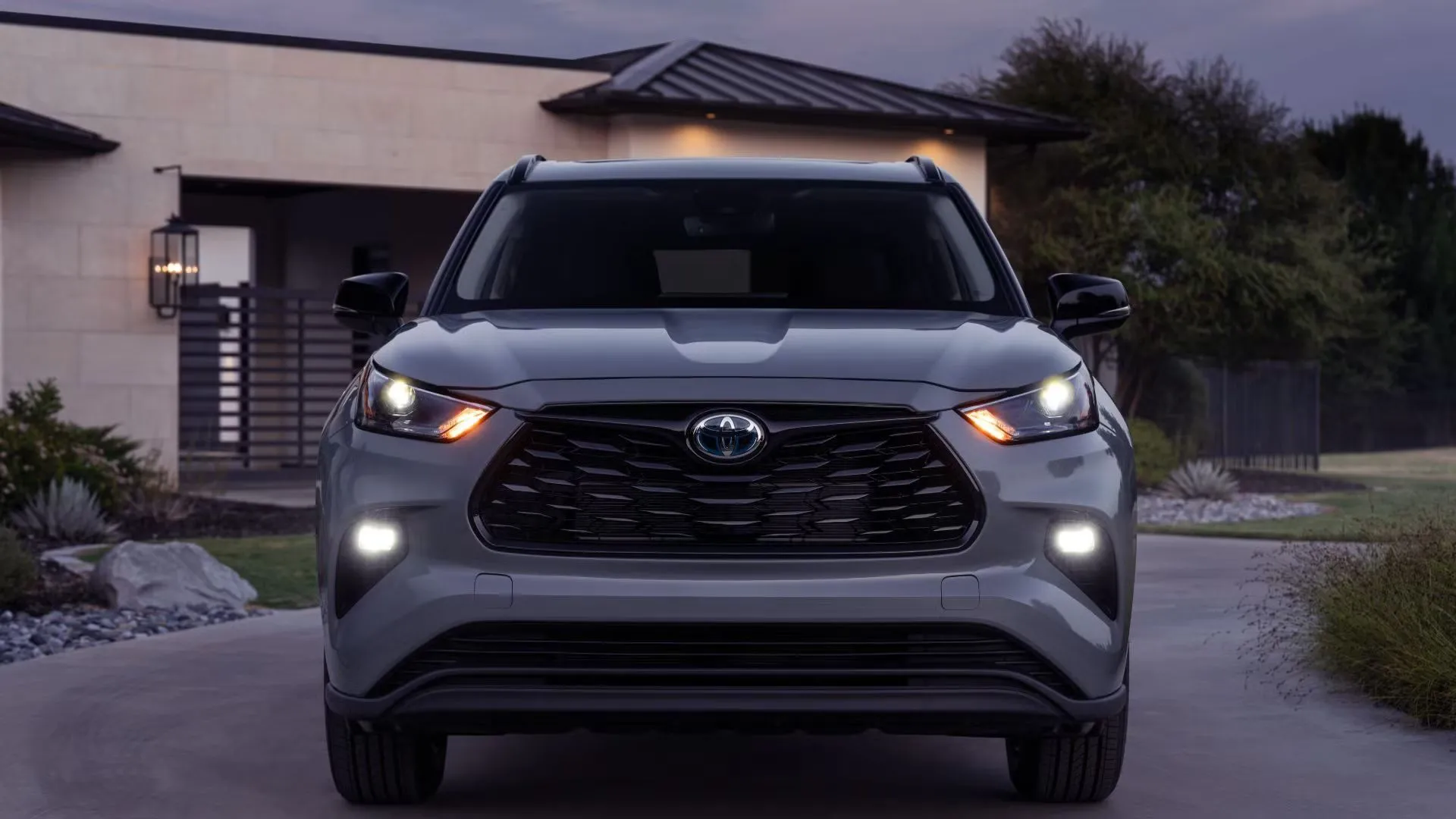 Góc phía trước của Toyota Highlander 2024 màu xám đỗ trên đường lái xe trước một ngôi nhà, đại diện cho SUV gia đình đáng tin cậy.