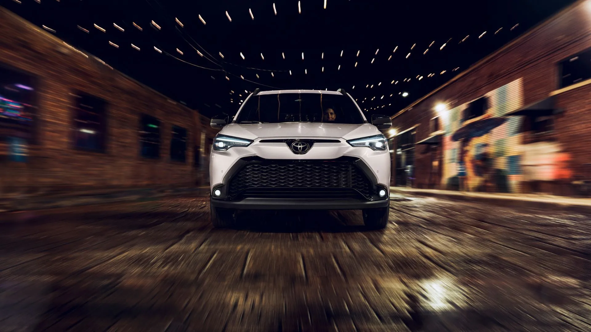 Hình ảnh động góc 3/4 phía trước của Toyota Corolla Cross Hybrid XSE 2025 màu trắng đang lái trên con phố lát đá trong thành phố vào ban đêm