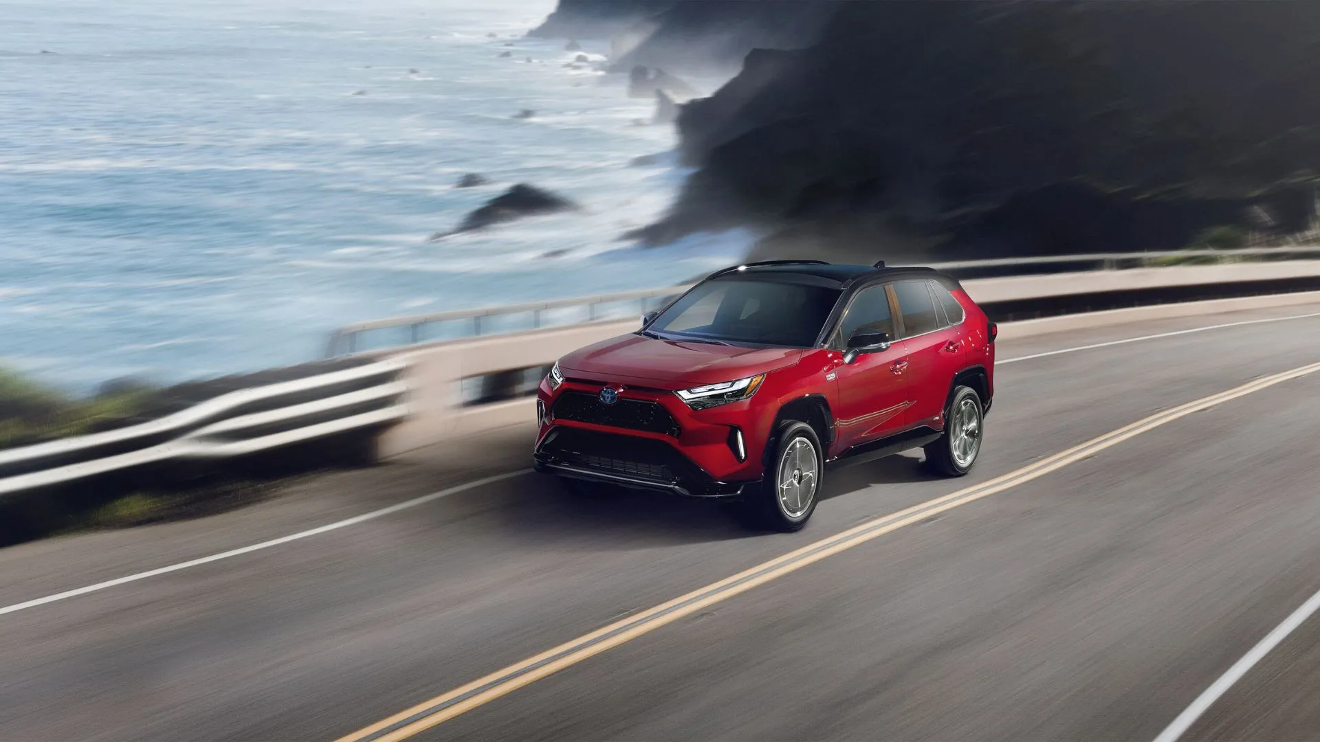 Hình ảnh động góc 3/4 phía trước của Toyota RAV4 Prime XSE 2024 màu đỏ đang chạy trên con đường ven biển, mẫu SUV hybrid plug-in hiệu quả.