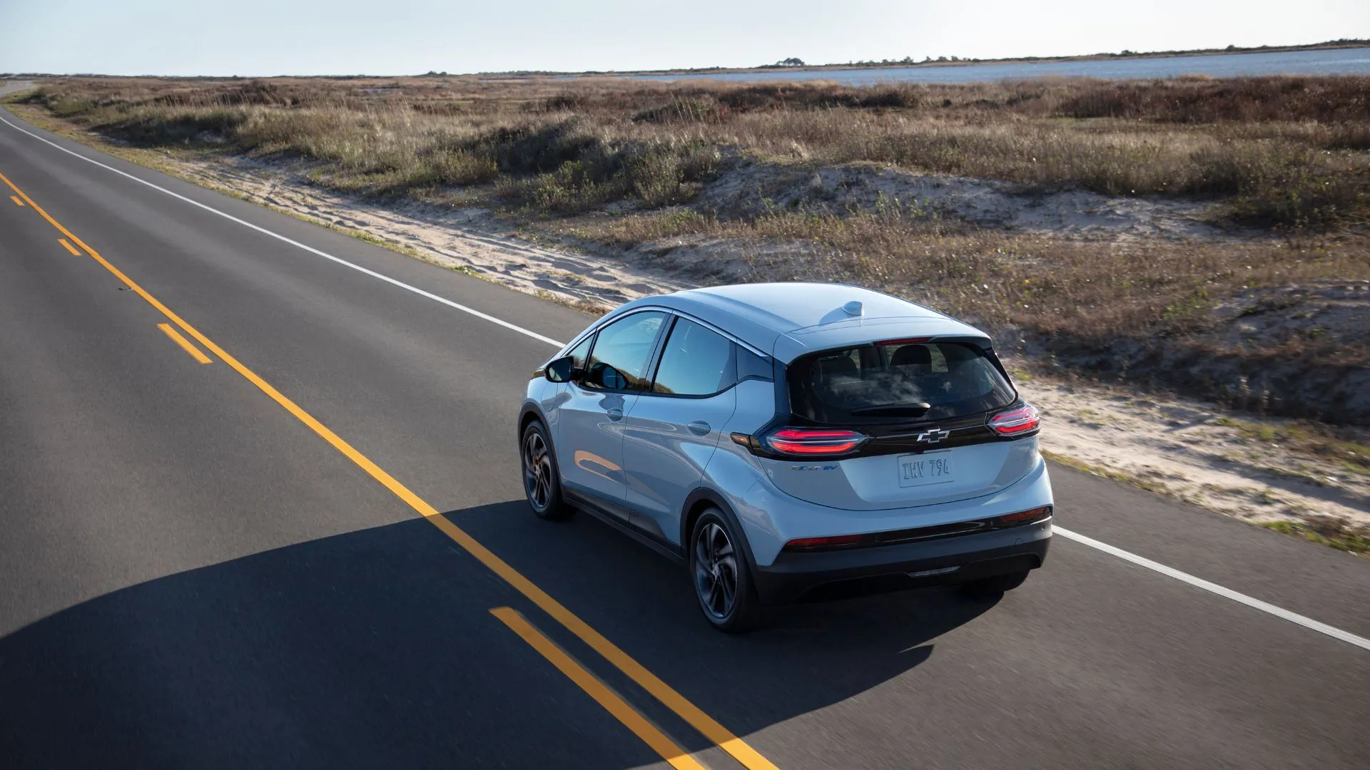Hình ảnh động xe Chevrolet Bolt EV 2022 màu bạc từ phía sau bên phải, chạy trên con đường nông thôn, cho thấy thiết kế nhỏ gọn và khả năng di chuyển linh hoạt của mẫu xe điện cũ này.