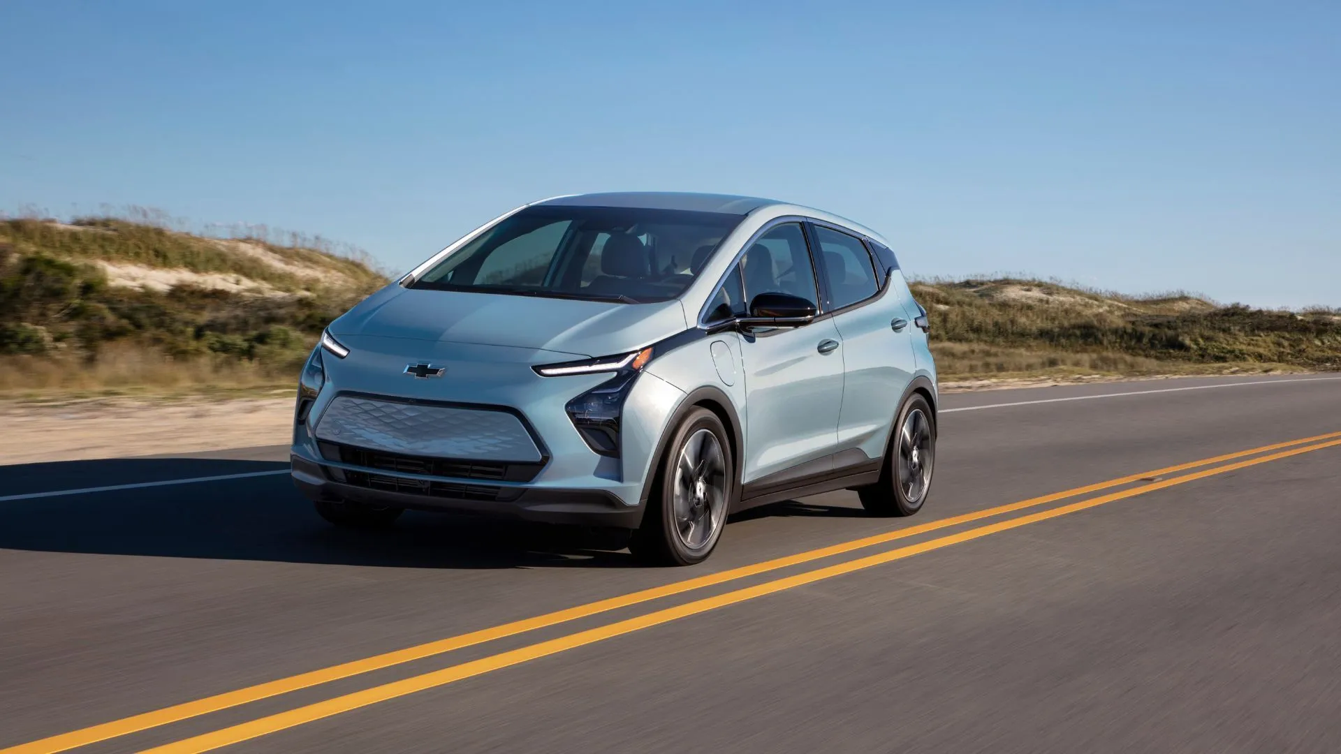 Hình ảnh động xe Chevrolet Bolt EV 2022 màu bạc từ phía trước bên phải, chạy trên con đường nông thôn, thể hiện sự bền bỉ và sẵn sàng vận hành của mẫu xe điện cũ đáng tin cậy.