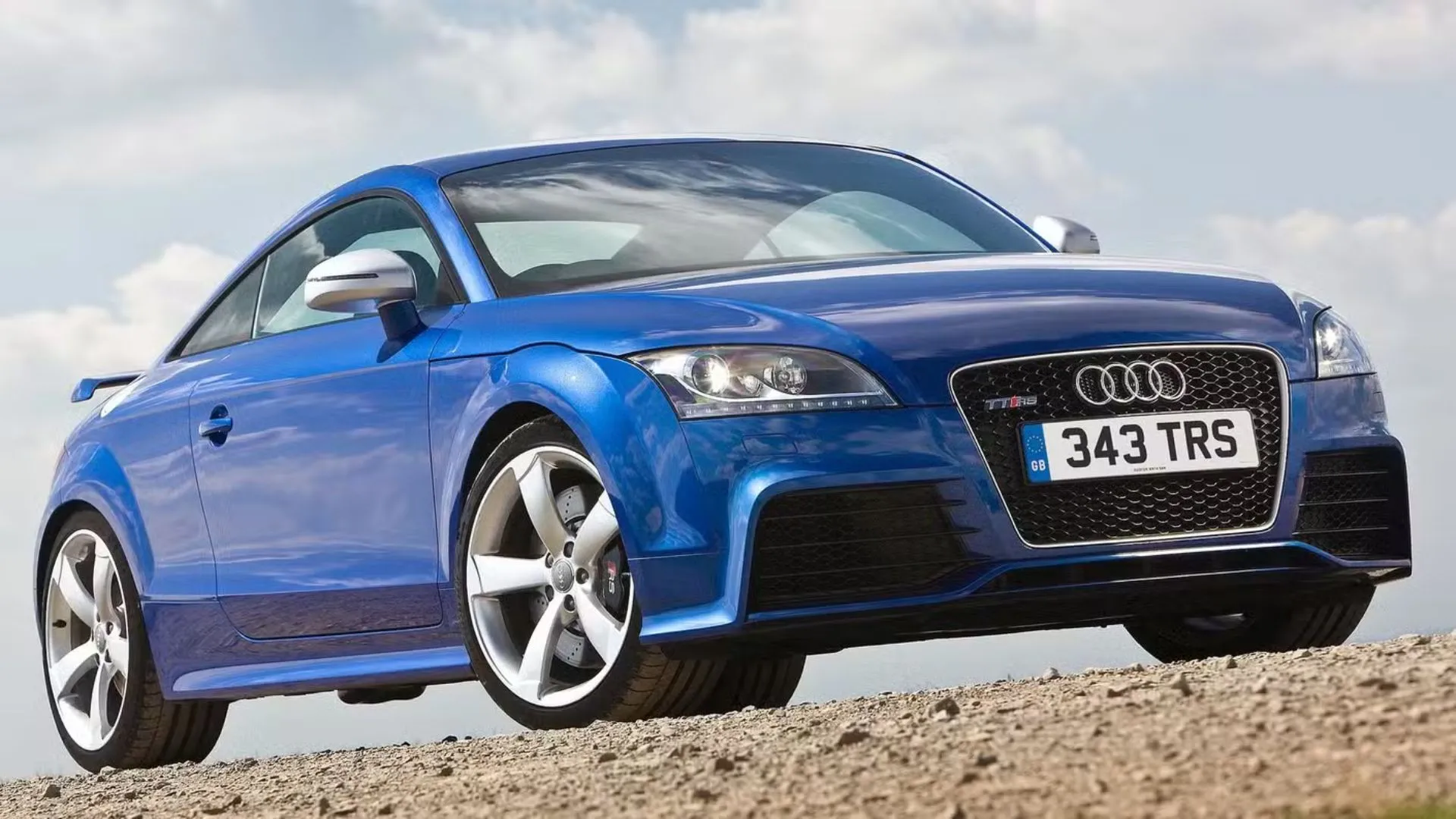 Hình ảnh góc 3/4 phía trước của chiếc Audi TT RS màu xanh dương đỗ trên nền sỏi, làm nổi bật thiết kế thể thao và mạnh mẽ