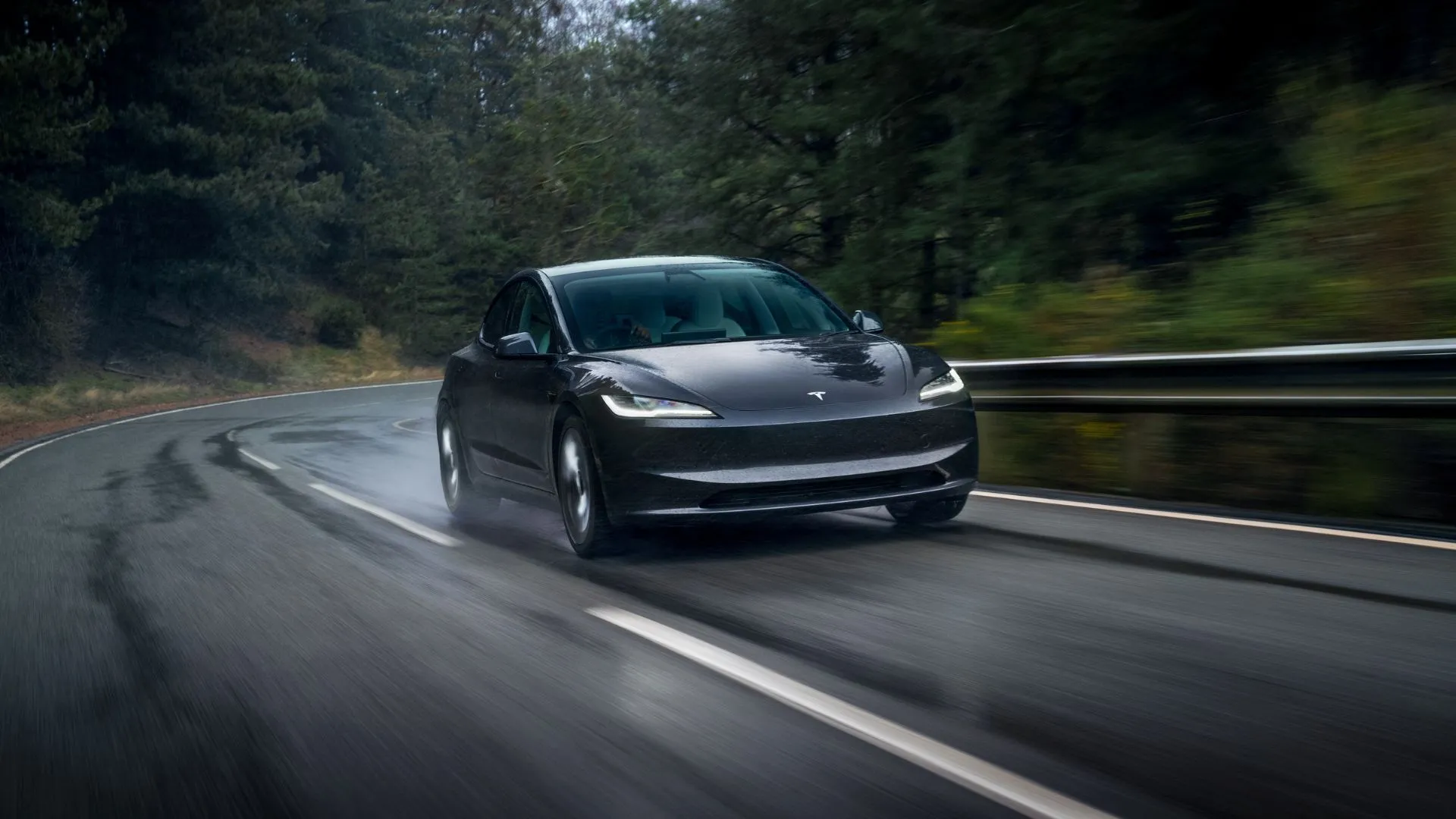Hình ảnh góc 3/4 phía trước của chiếc Tesla Model 3 2024 màu xám đang chạy trên con đường nông thôn ướt át.