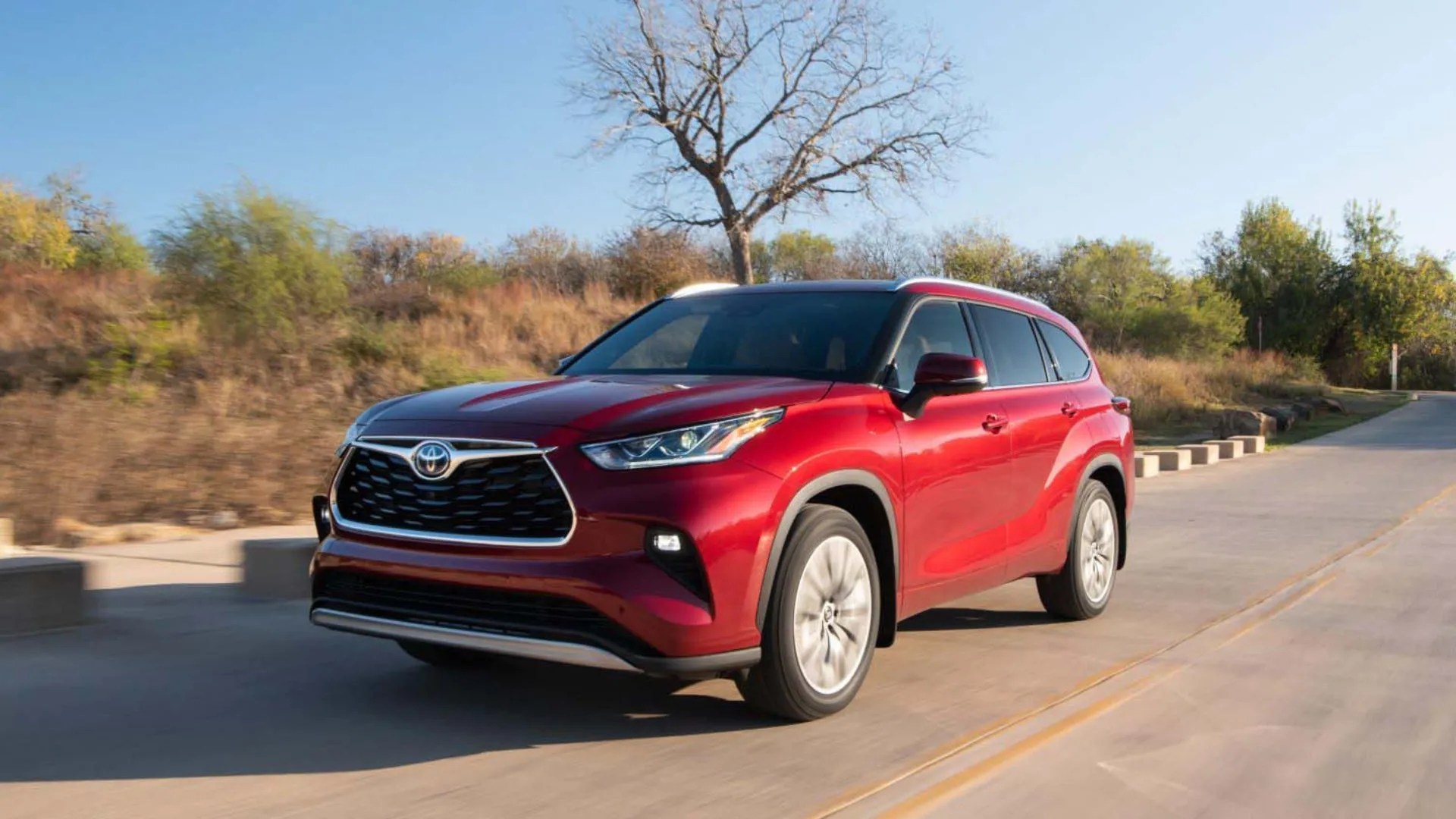 Hình ảnh mặt trước ba phần tư của một chiếc Toyota Highlander Platinum Hybrid 2024 màu đỏ đang lái trên con đường nông thôn.