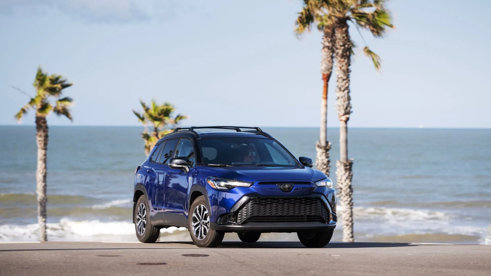 Hình ảnh tĩnh góc 3/4 phía trước của Toyota Corolla Cross Hybrid SE 2024 màu xanh với cây cọ và biển làm nền