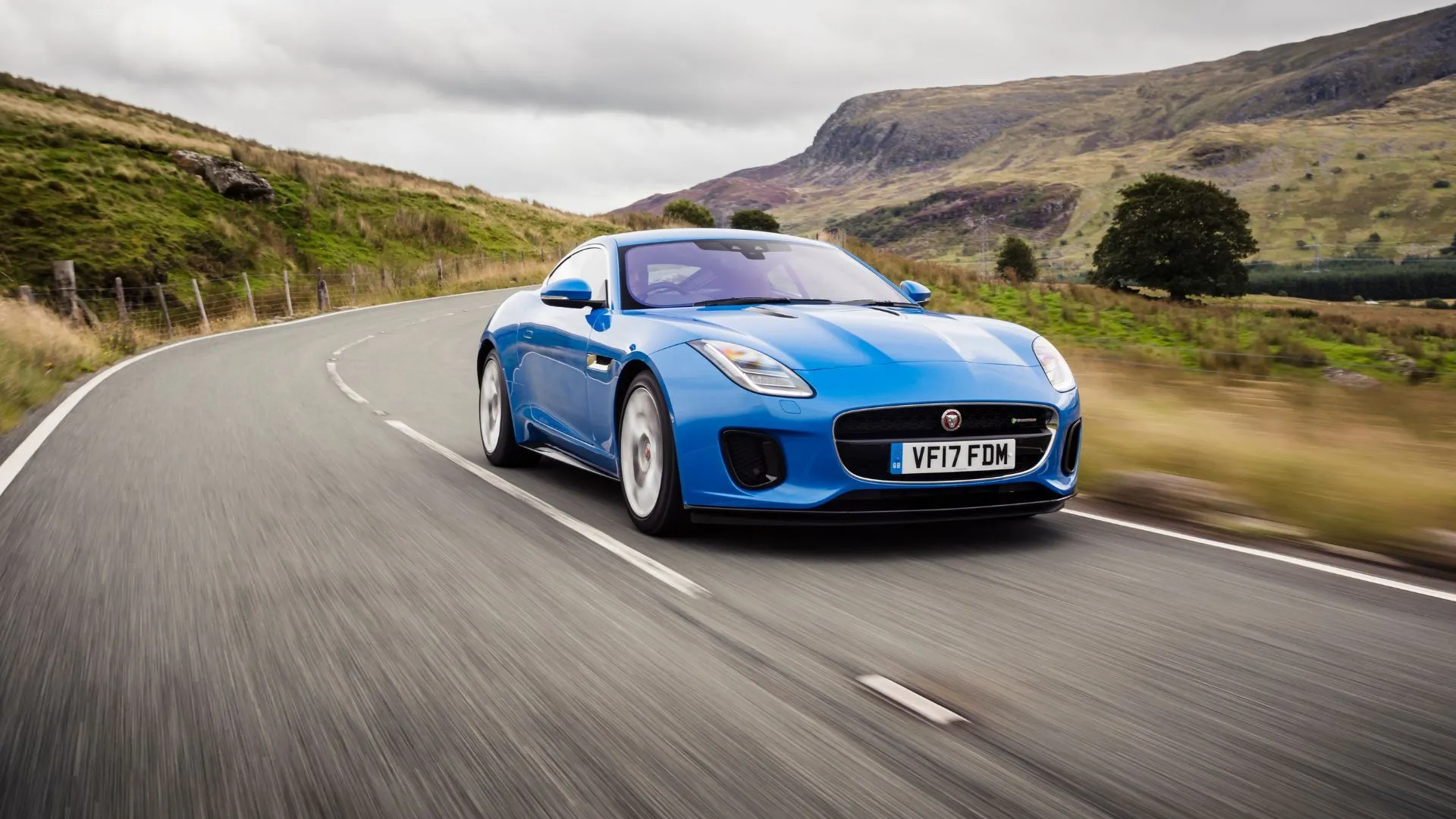 Jaguar F-TYPE R-Dynamic Coupe màu xanh dương đời 2017 lái trên đường nông thôn uốn lượn, thiết kế quyến rũ và âm thanh ống xả đặc trưng.