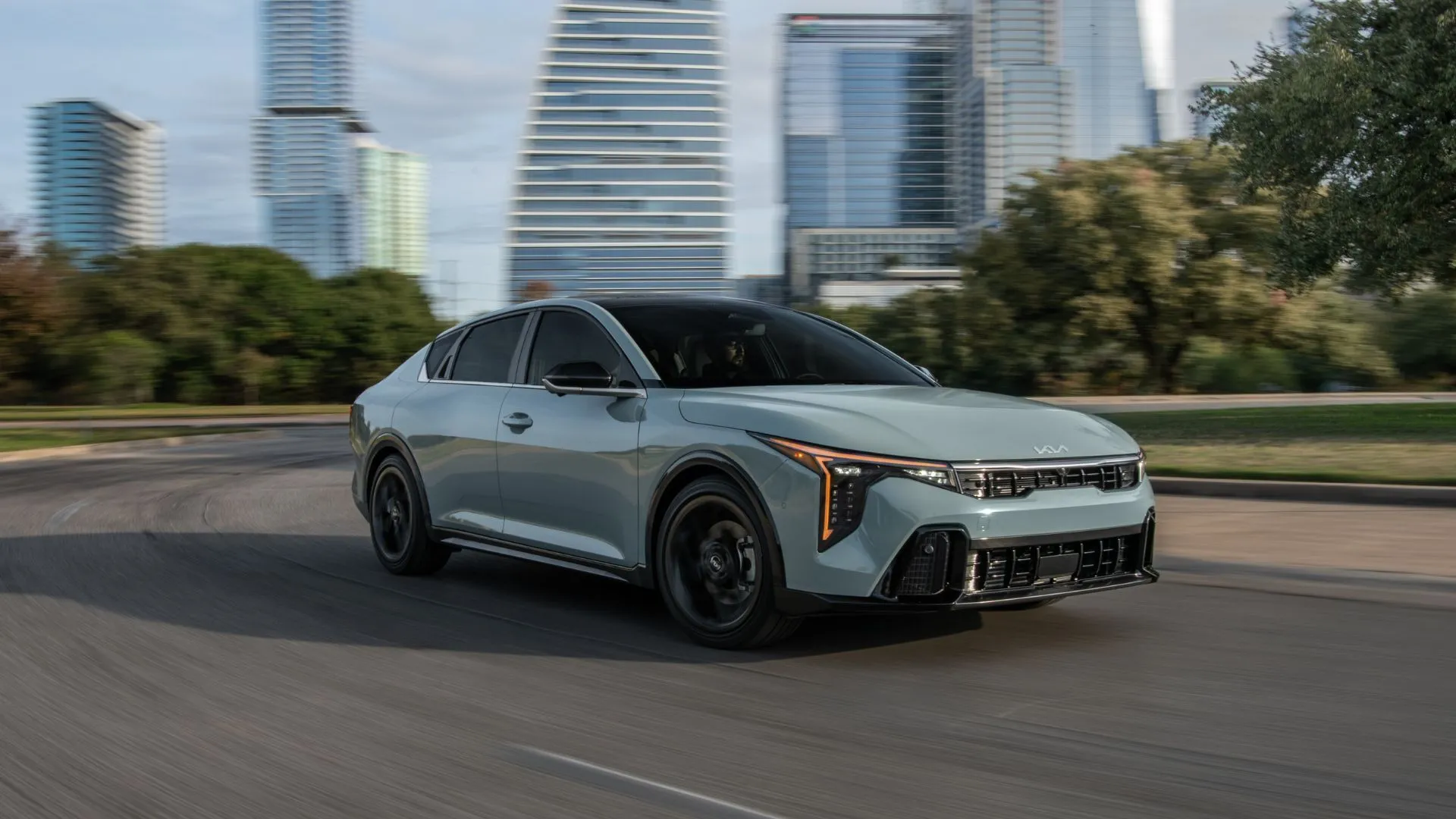 Kia K4 2025 màu xám đang di chuyển qua khu đô thị với kiến trúc hiện đại