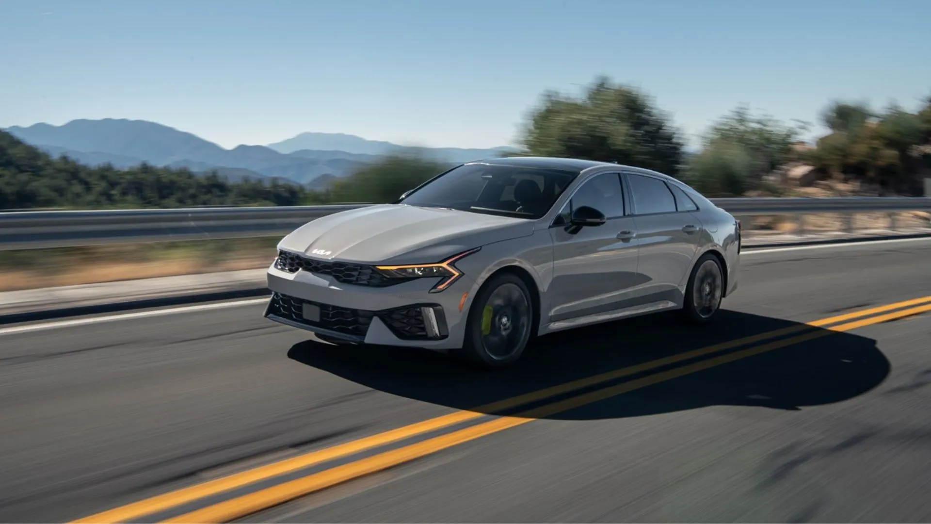 Kia K5 2025 màu xám đang lái trên con đường nông thôn với núi non phía sau.