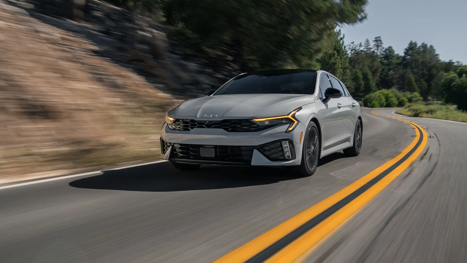 Kia K5 GT 2025 màu xám bạc lướt trên đường núi, thể hiện sự năng động