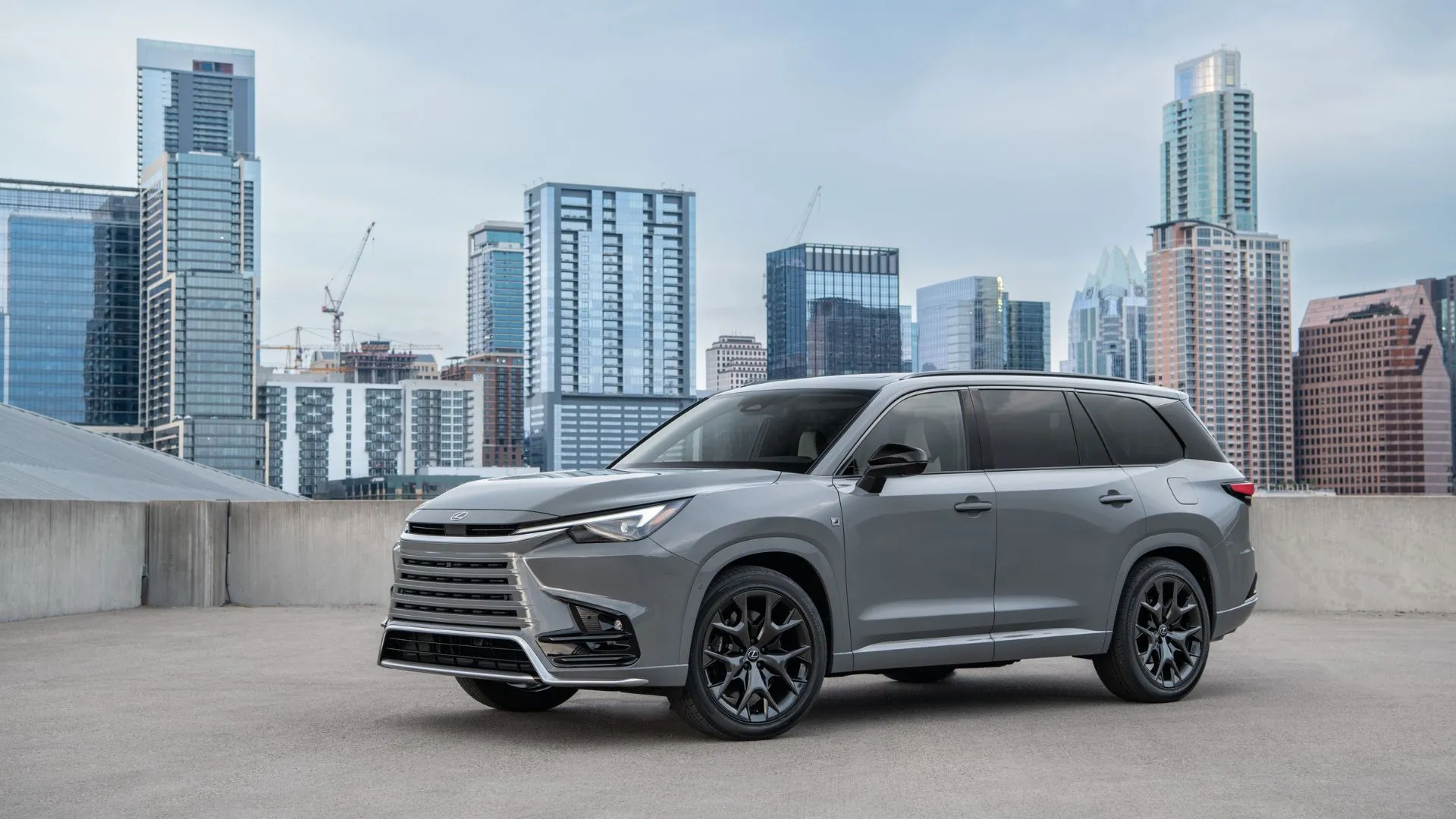Lexus TX 500h F Sport Performance 2024 màu xám đang đỗ trên sân thượng với các tòa nhà chọc trời phía sau, toát lên vẻ thể thao và sang trọng.