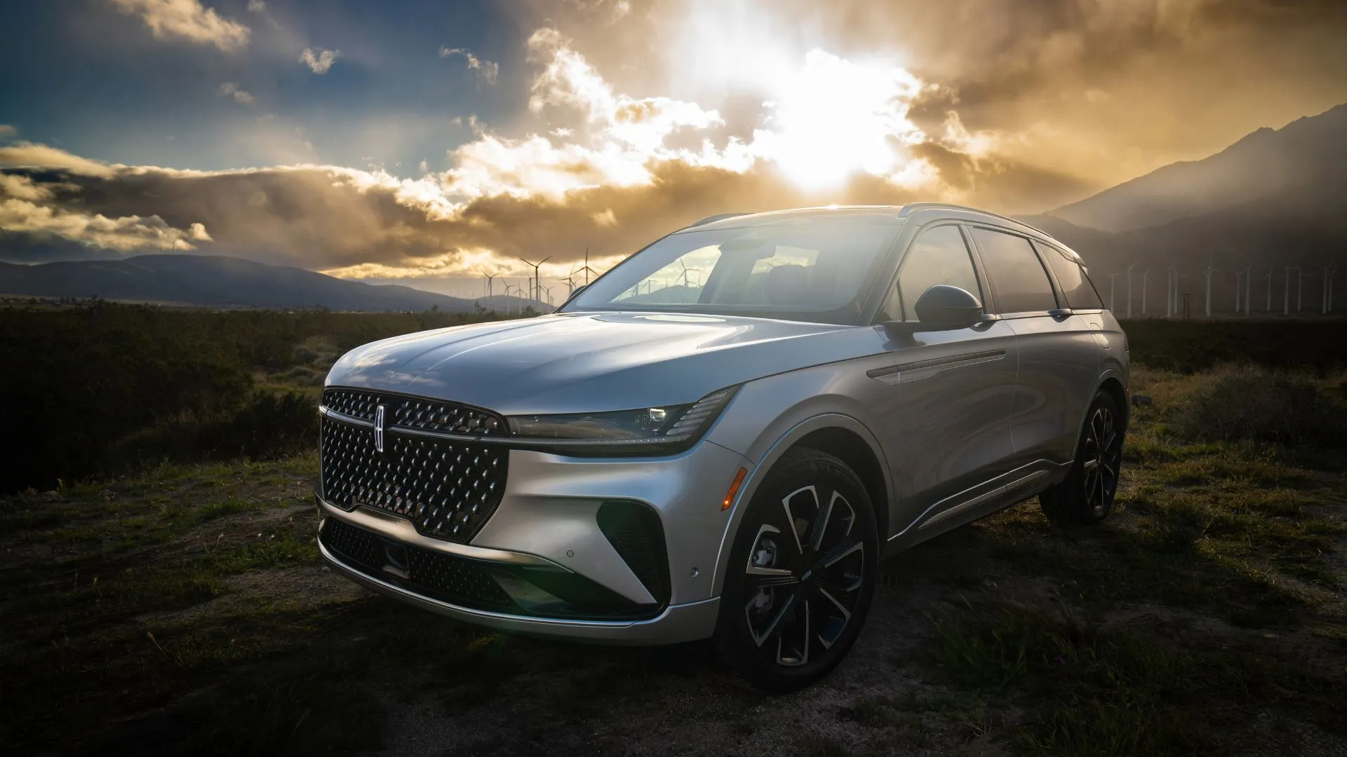 Lincoln Nautilus Reserve Hybrid 2024 màu bạc vào lúc hoàng hôn với các tuabin gió phía sau, thể hiện sự kết hợp hài hòa giữa công nghệ và hiệu quả.