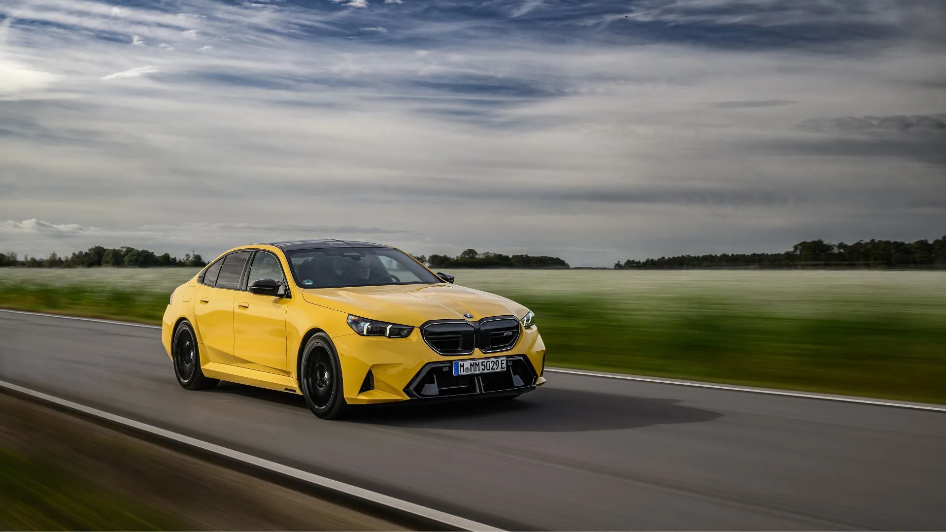 Mẫu sedan BMW M5 2025 màu vàng năng động nhìn từ góc 3/4 phía trước, đang di chuyển trên con đường nông thôn với những cánh đồng xanh mướt phía sau.