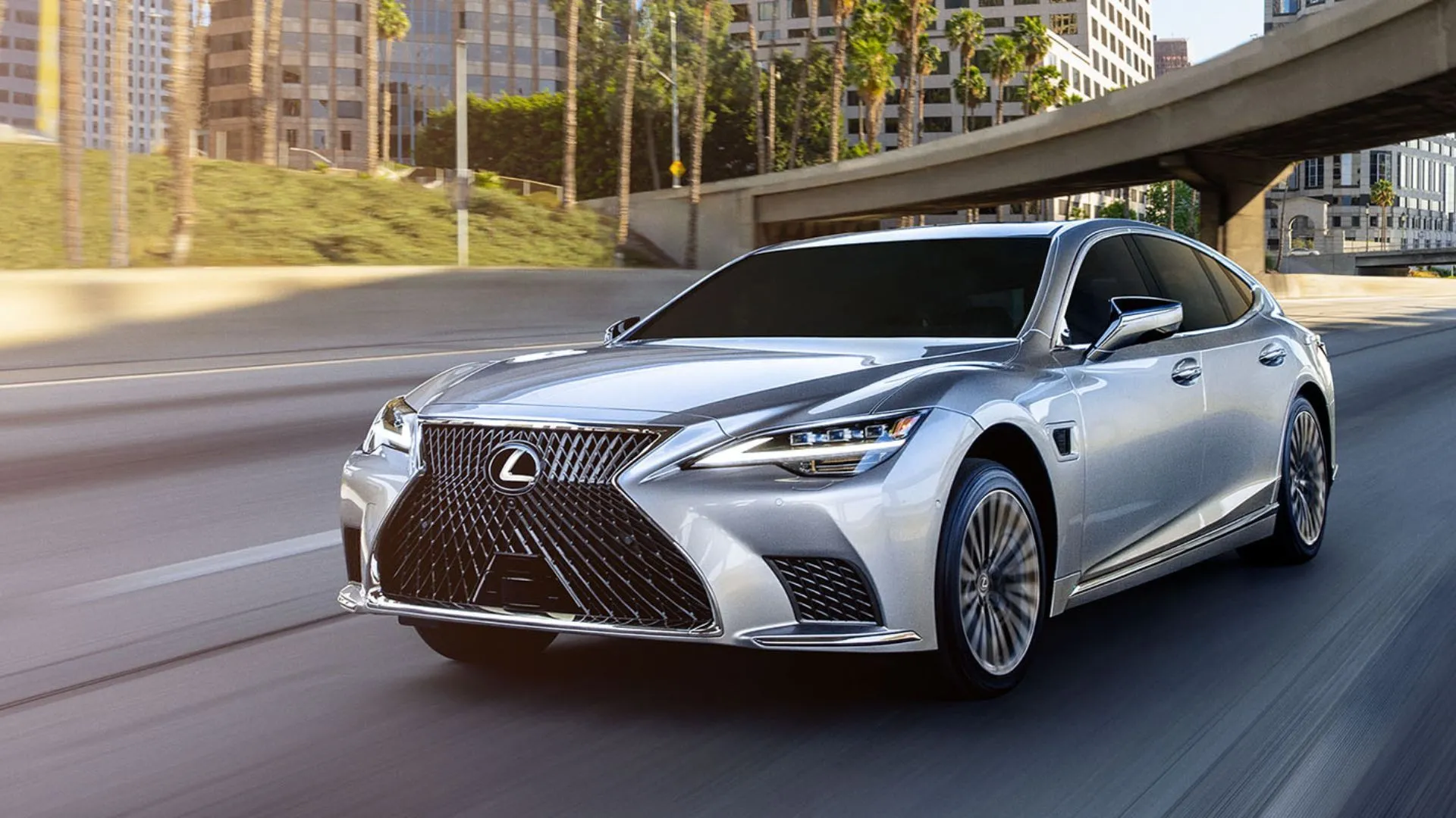 Mẫu sedan Lexus LS 500h 2025 màu bạc nhìn từ góc 3/4 phía trước, đang di chuyển qua thành phố, toát lên vẻ thanh lịch và hiện đại.