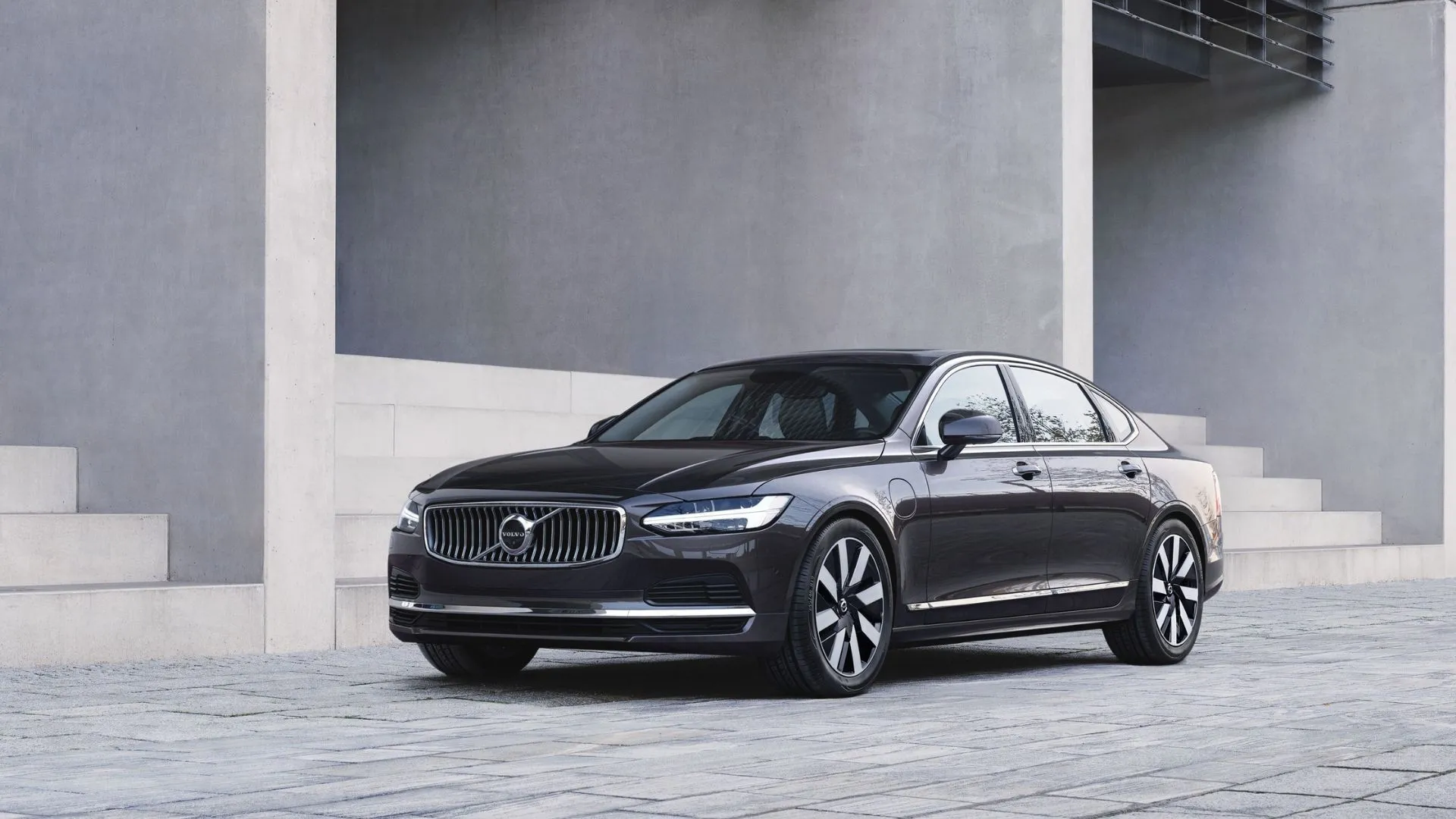 Mẫu sedan Volvo S90 Recharge T8 AWD 2024 màu xám nhìn từ góc 3/4 phía trước, đang đỗ cạnh tòa nhà với những bậc thang bên ngoài, toát lên vẻ hiện đại và thanh lịch.