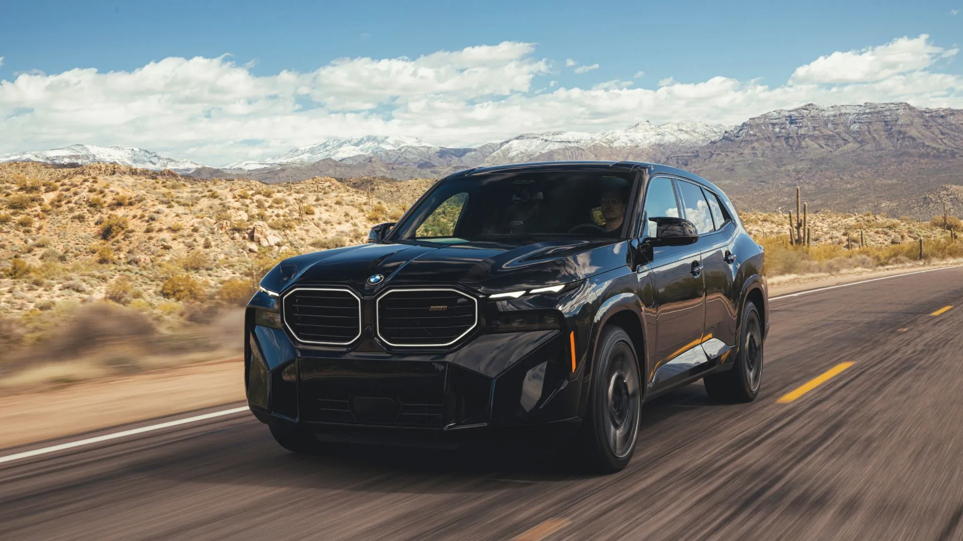 Mẫu SUV BMW XM 2023 màu đen năng động nhìn từ góc 3/4 phía trước, đang di chuyển trên con đường nông thôn với những ngọn núi hùng vĩ phía sau.