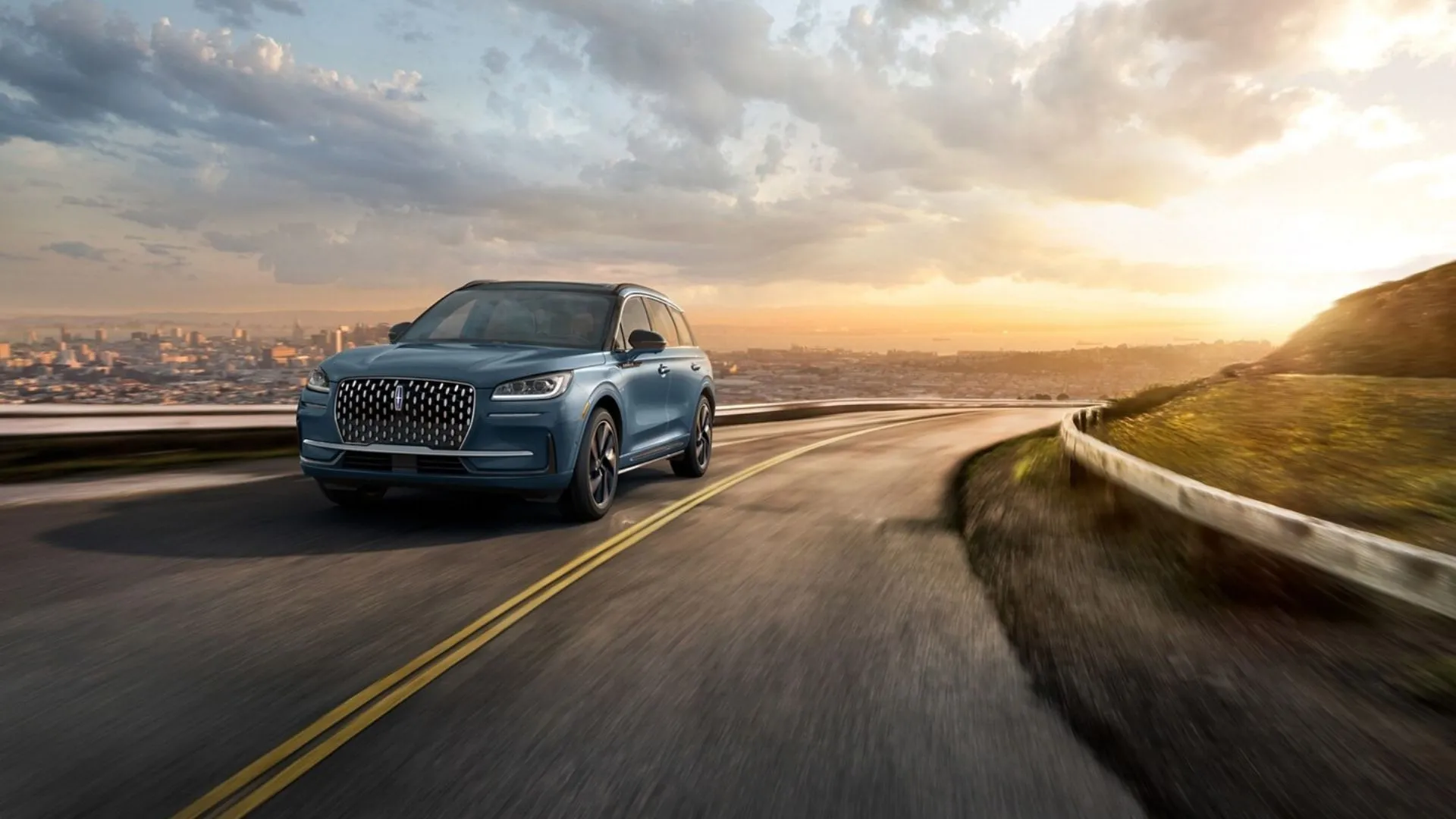 Mẫu SUV Lincoln Corsair Grand Touring 2025 màu xanh năng động nhìn từ góc 3/4 phía trước, đang di chuyển trên con đường quanh co với cảnh quan thành phố ở hậu cảnh.