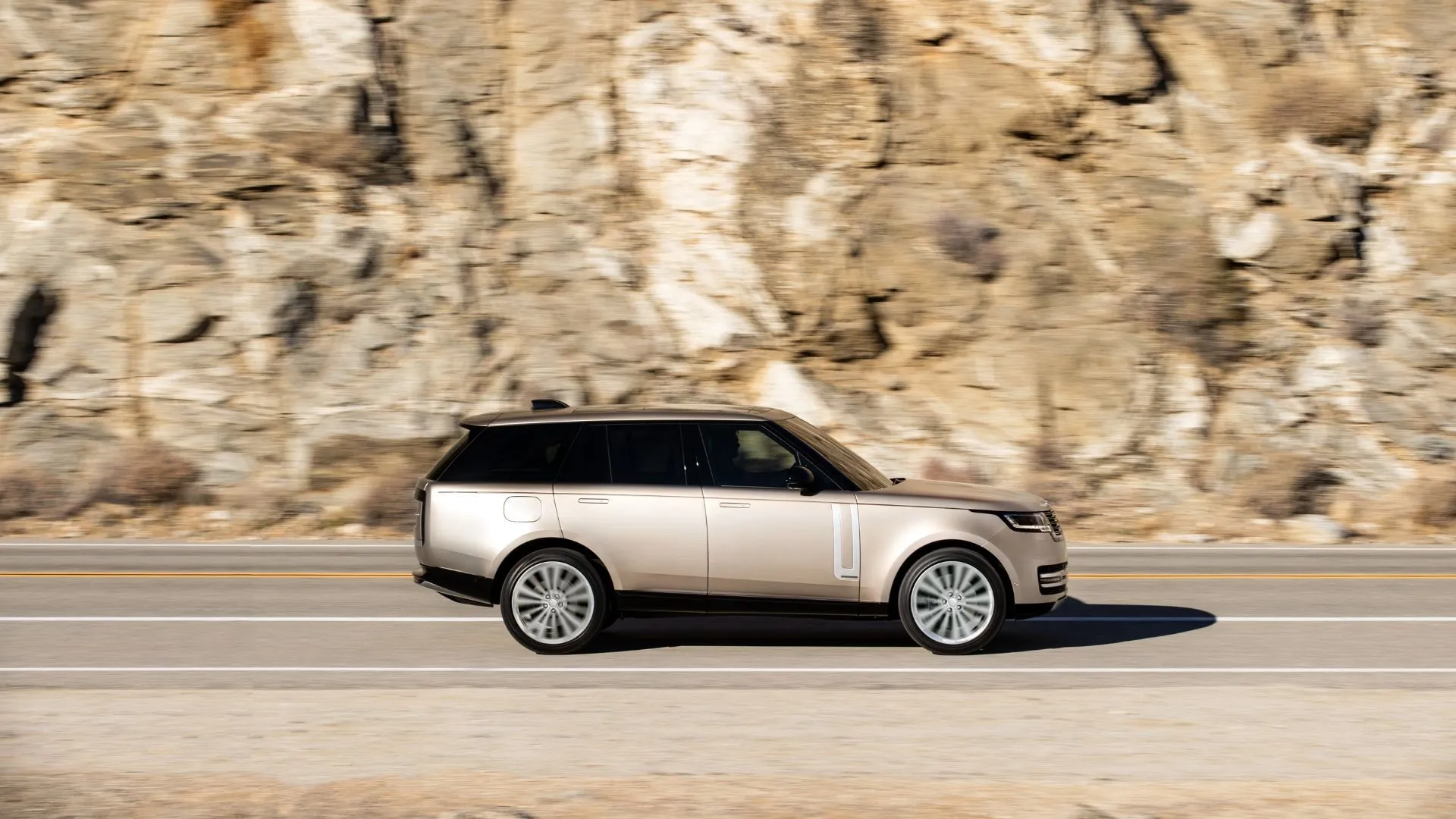 Mẫu SUV Range Rover 2022 màu vàng năng động nhìn từ bên hông, đang di chuyển trên con đường nông thôn với vách đá sừng sững phía sau.