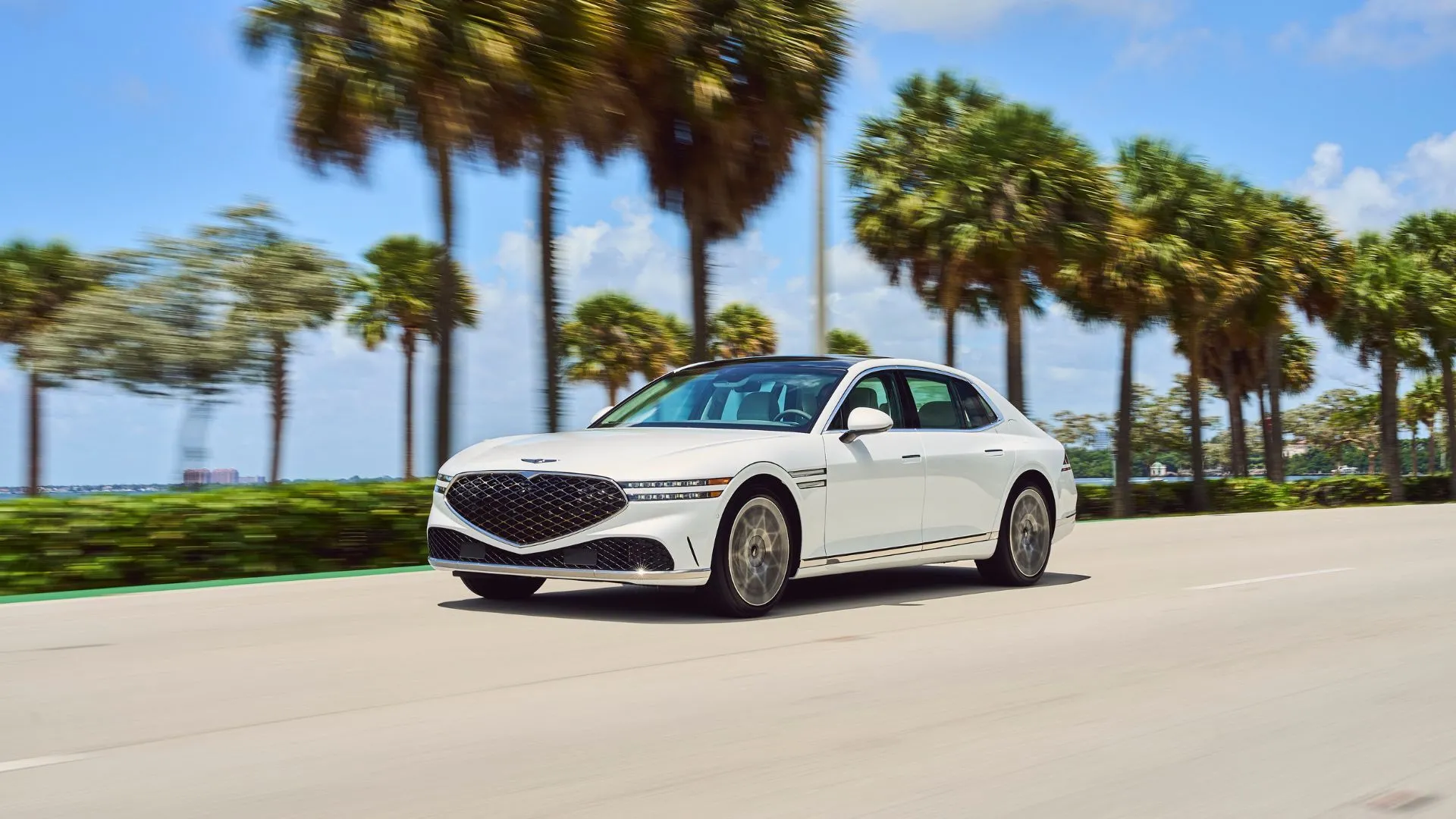 Mẫu xe Genesis G90 2024 màu trắng năng động nhìn từ góc 3/4 phía trước, đang di chuyển trên đường cao tốc với hàng cọ xanh mát phía sau.