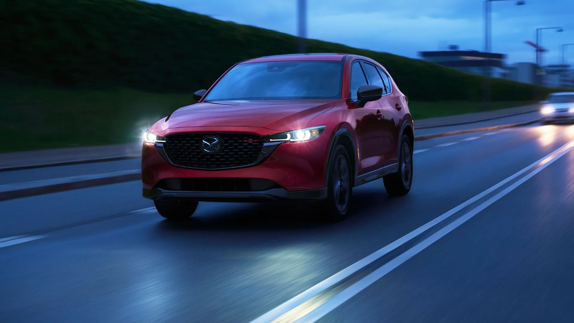 Mazda CX-5 2025 màu đỏ bóng loáng đang di chuyển trên đường phố vào ban đêm với ánh đèn thành phố rực rỡ phía sau.