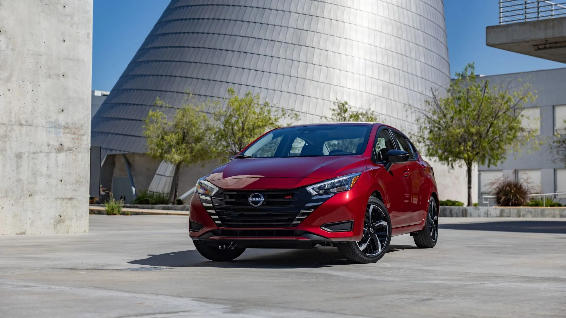 Sedan Nissan Versa 2025 màu đỏ đỗ trước tòa nhà kính hiện đại, minh họa cho sự hồi phục doanh số của Nissan