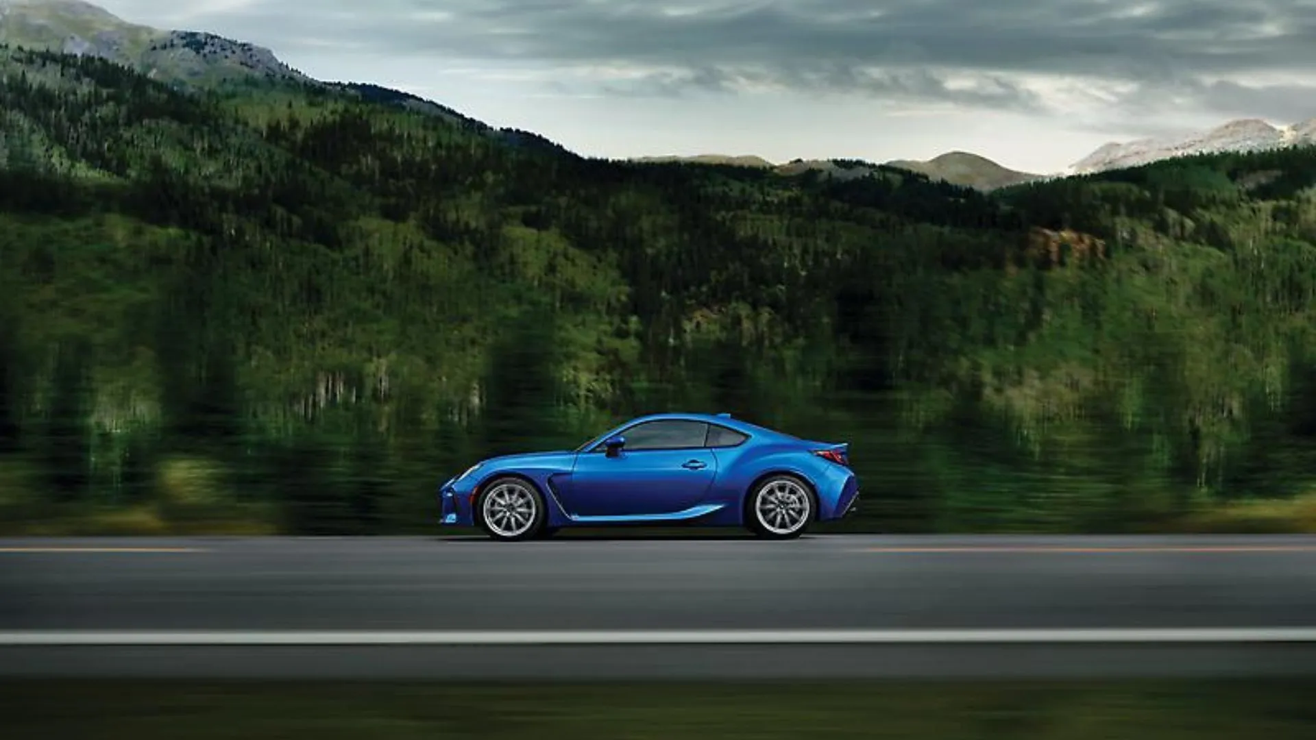 Subaru BRZ 2024 màu xanh dương trên đường nông thôn với núi và cây cối phía sau, nhìn ngang