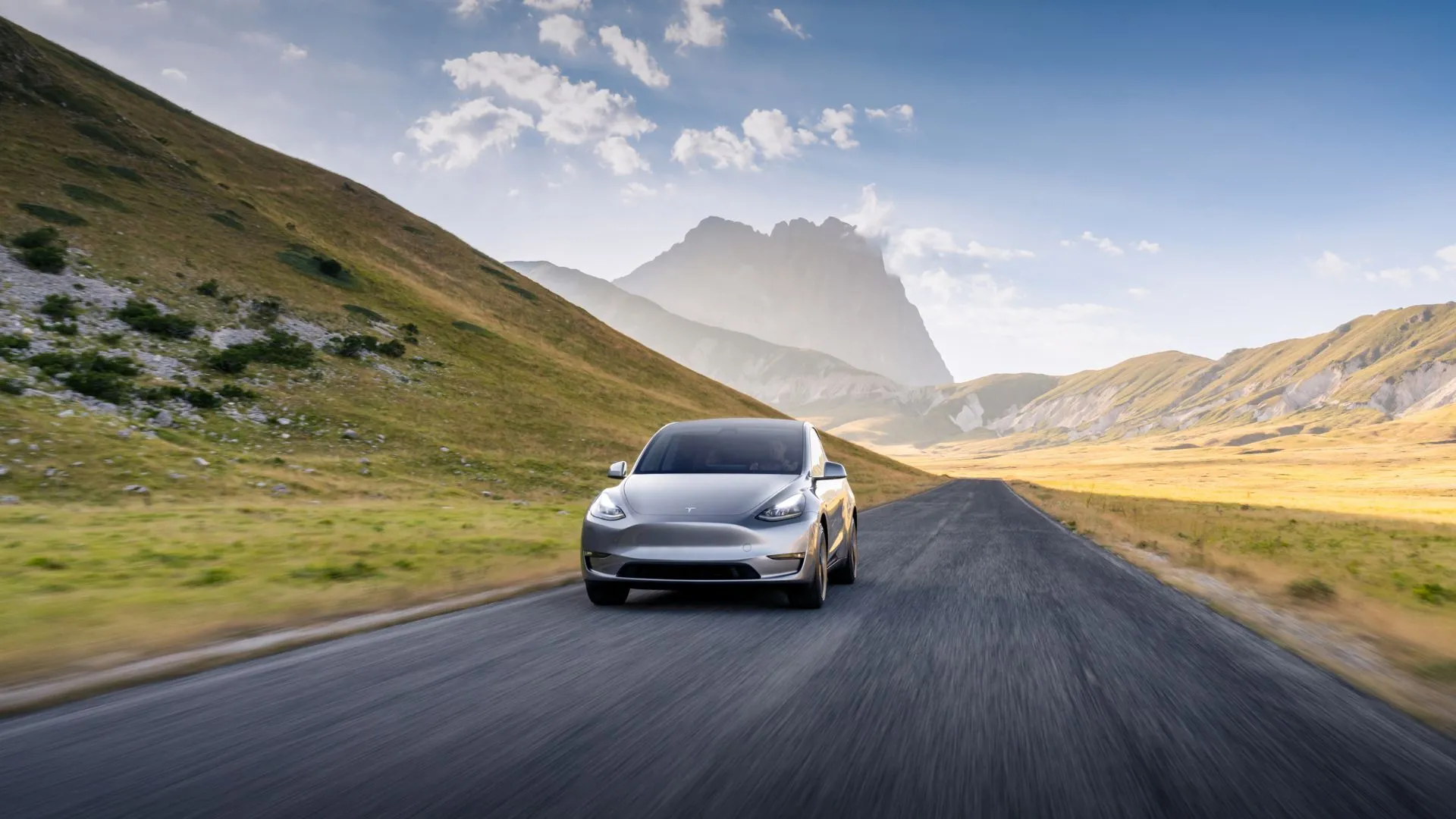 Tesla Model Y 2023 màu bạc lăn bánh trên đường quê với cảnh núi non hùng vĩ, thể hiện trải nghiệm lái xe điện tiện nghi và đẳng cấp.