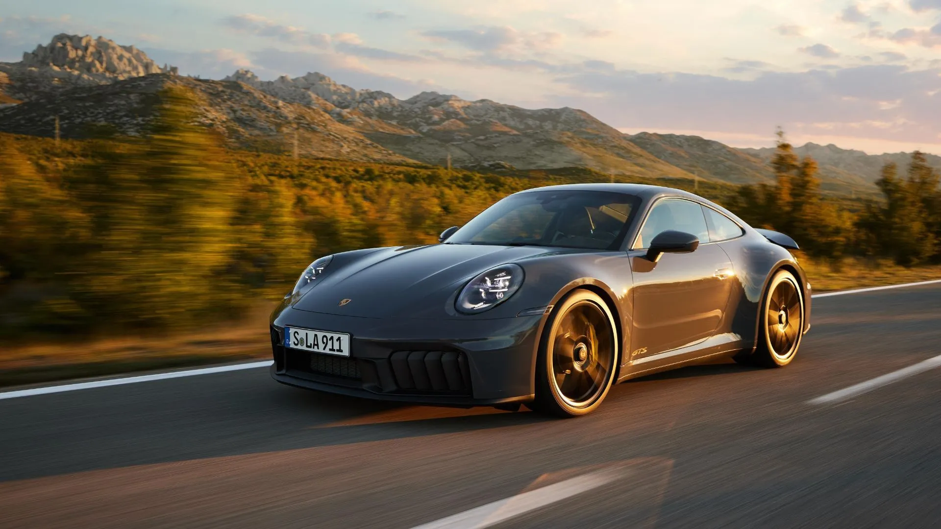 Toàn cảnh chiếc Porsche 911 Carrera GTS Hybrid 2024 màu xám đang chạy trên đường nông thôn