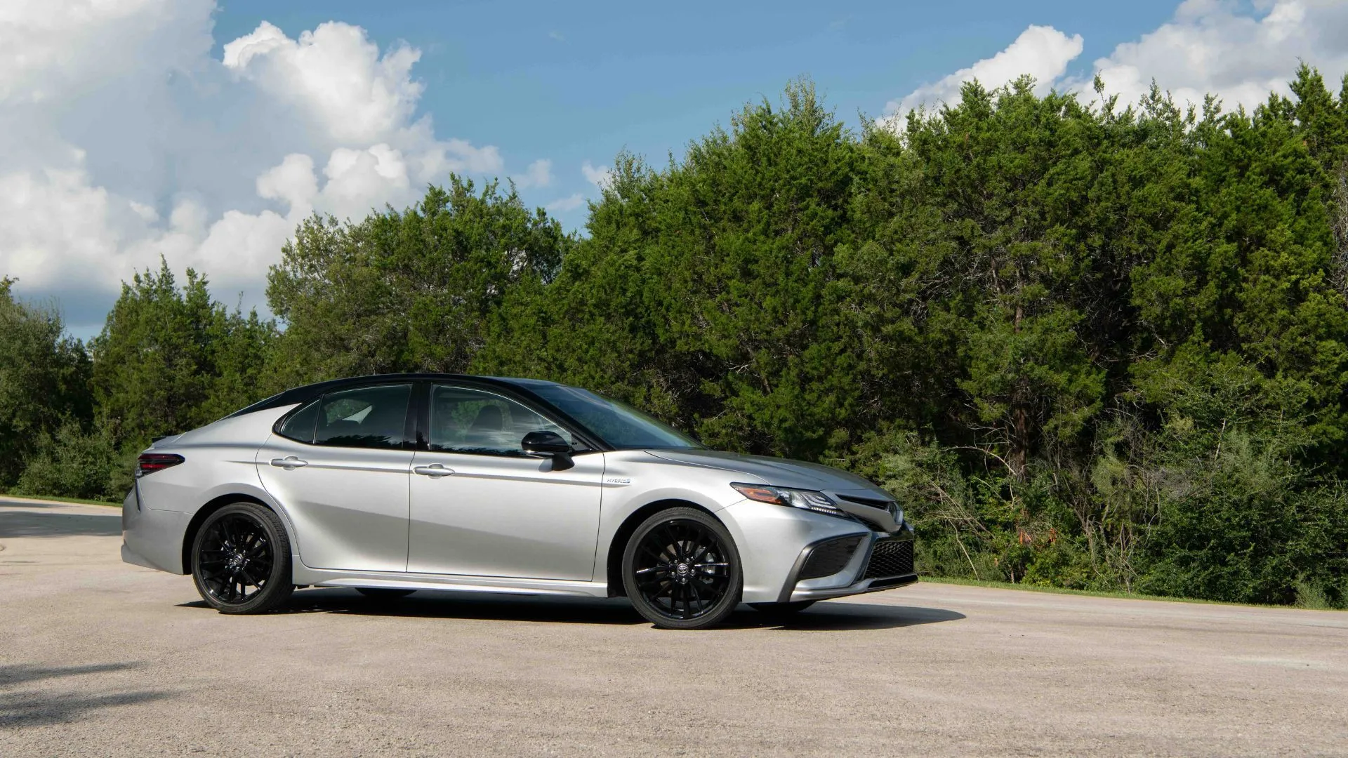 Toyota Camry Hybrid 2022 XSE màu bạc đỗ trên đường nhựa với hàng cây phía sau, tôn lên vẻ ngoài tinh tế và thiết kế sedan hiện đại.